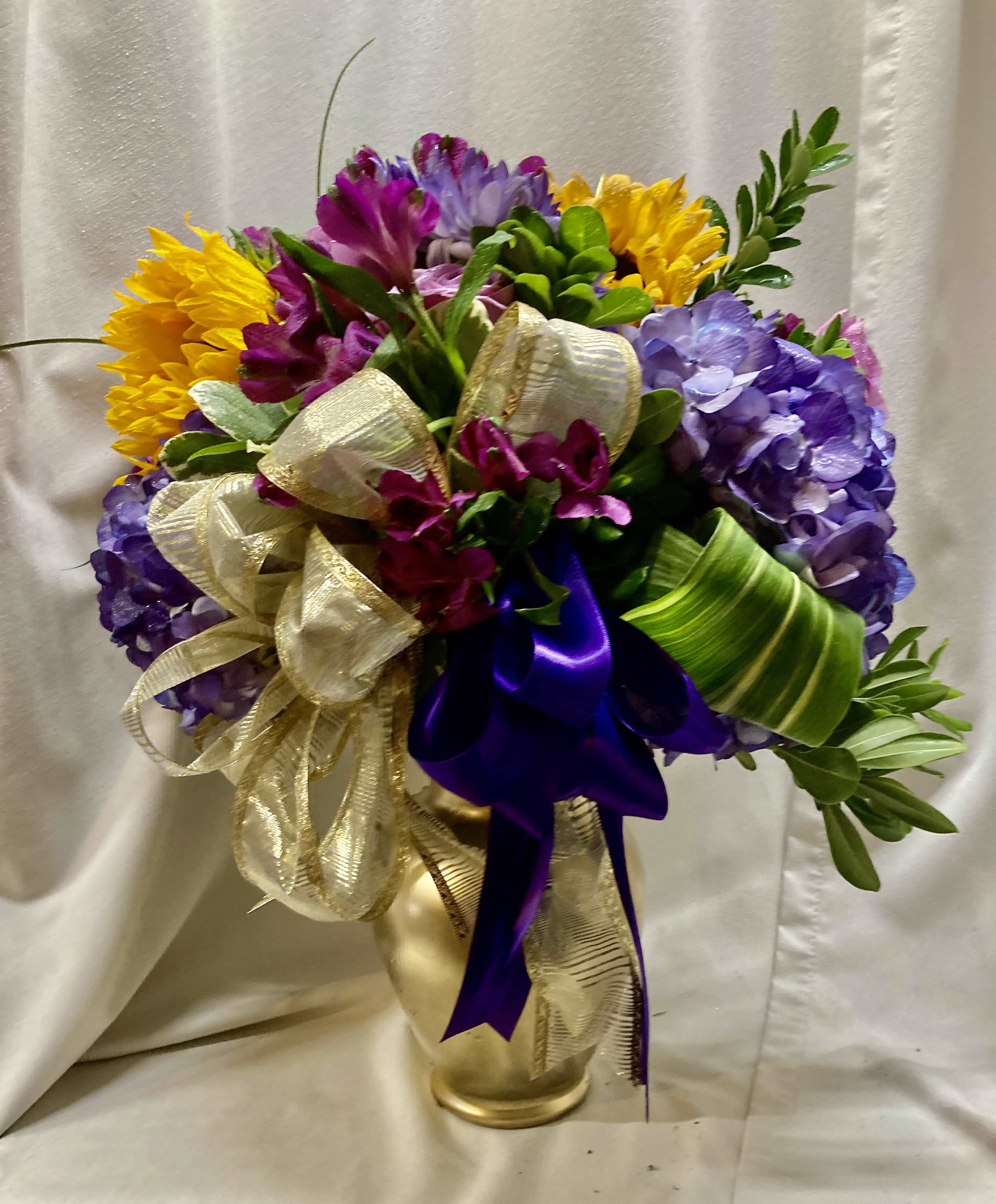 ENLIGHTENED - A bold celebration of color, this flower arrangement pairs lush purple hydrangeas with bright yellow blooms and magenta alstroemeria for an energetic birthday bouquet or congratulations gift that reads joyful from across the room. Designed to make the recipient feel truly celebrated, ENLIGHTENED brings a luxurious, playful mood-perfect for milestone birthdays, promotions, or any occasion that calls for a festive flourish.  Dense, rounded clusters of purple hydrangea form the visual heart, while vivid yellow flowers and magenta alstroemeria add lively contrast and texture. The composition is finished with large metallic gold, purple, and green ribbons and sits in a gleaming gold vase, creating a striking contrast that elevates any tabletop or mantle. Sized to impress without overwhelming, it's a decorative gift arrangement that delivers high-impact color and presence.  Give this to someone you want to see smile-she'll feel noticed and celebrated the moment it arrives. For best enjoyment, keep the vase filled with fresh water and place away from direct heat or sunlight; fresh flower care will help sustain the vibrant look through the celebration.