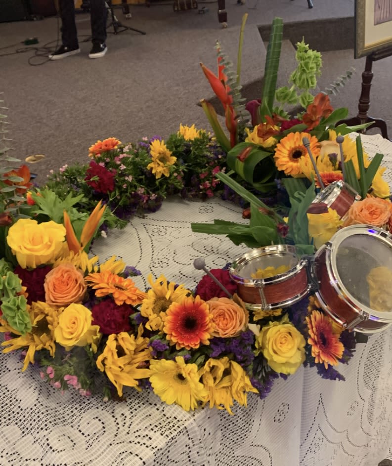 DRUMMERS URN - A vivid circular wreath of celebration, this flower arrangement is perfect as a ceremonial centerpiece or a festive sympathy tribute. The drum-themed design-small decorative drums nested within the floral ring-gives the piece a musical, commemorative spirit that suits memorial services, cultural ceremonies, or any event where you want the recipient to feel honored and remembered.  Bright yellow and orange roses and gerbera daisies form the dominant halo, their sunlit tones balanced by pockets of pink, red, and purple blooms for depth and contrast. Carnations and assorted filler flowers create a dense, handcrafted texture around the ring; the arrangement is displayed flat on a lace-covered table for an elegant, traditional presentation. The combination of warm hues and lively shapes reads as both joyful and respectful.  Designed to celebrate a life, mark a milestone, or add a meaningful focal point to an event, this wreath sends a message of warmth and remembrance to the person you're honoring. Because it's arranged flat as a circular wreath, it's ready to lay on a table or stand as directed by your venue; please let us know any delivery details so we can position it properly for the occasion.