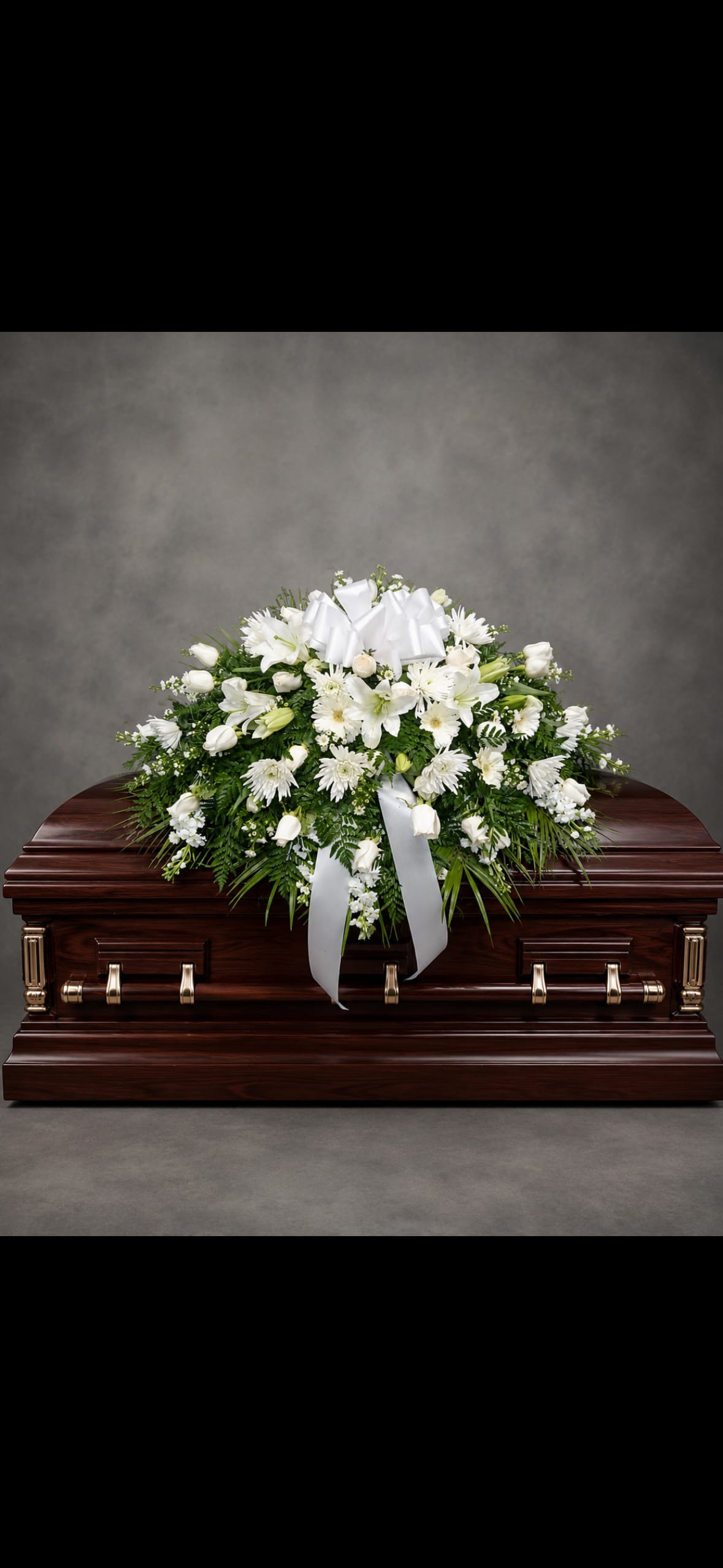 White Casket Spray - A serene, traditional white casket spray that conveys deep sympathy with quiet elegance-designed to rest across the top of a dark wood casket for a funeral or memorial service. The arrangement brings together white roses, white lilies, chrysanthemums, carnations, and daisy-like blooms to create a full, rounded composition that reads reverent and soothing for the person you're honoring.  Abundant green foliage frames the creamy whites and adds soft texture while a wide white satin bow crowns the spray; ribbon tails drape down the front for a formal, finished presentation. Sized to span the length of a casket, this design is crafted for visibility from the front and sides and arranged to maintain its shape throughout the service.  A meaningful way to express sympathy and respect, this spray offers a timeless look that comforts mourners and honors the deceased. Care is handled by the florist for delivery and placement on the casket; please confirm service time and cemetery or funeral home details when ordering so the arrangement arrives for the ceremony.