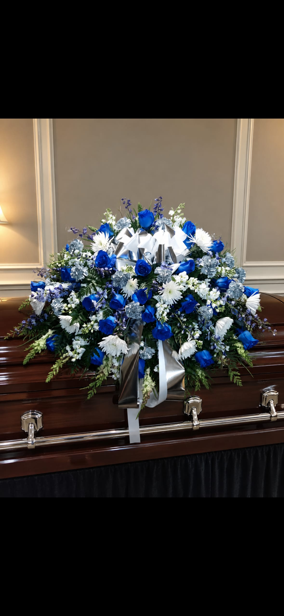 Blue And White Casket Spray - A dignified blue-and-white casket spray that offers a cool-toned, respectful tribute for a funeral or memorial service. Designed to rest across the top of a casket, this funeral arrangement combines roses and chrysanthemum-like blooms in blue, white, and green hues so the person you're honoring feels genuinely remembered and celebrated. Silver and white satin ribbon accents at the center add a quiet, formal touch that reads as both comforting and elegant.  Dense clusters of roses, chrysanthemums and delicate filler flowers create a full, slightly cascading composition that drapes gracefully down the sides of the casket. The spray is arranged broadly to provide a prominent service display; satin ribbon gathers at the center and trails downward for a finished, ceremonial look. This is the kind of floral tribute that conveys sympathy and respect with timeless restraint.  Appropriate for immediate family, close friends, or as a memorial centerpiece, this casket spray sends a clear message of honor and remembrance. If you need guidance on sizing or delivery to a funeral home, let us help coordinate placement and timing to ensure the tribute arrives fresh and ready for the service.