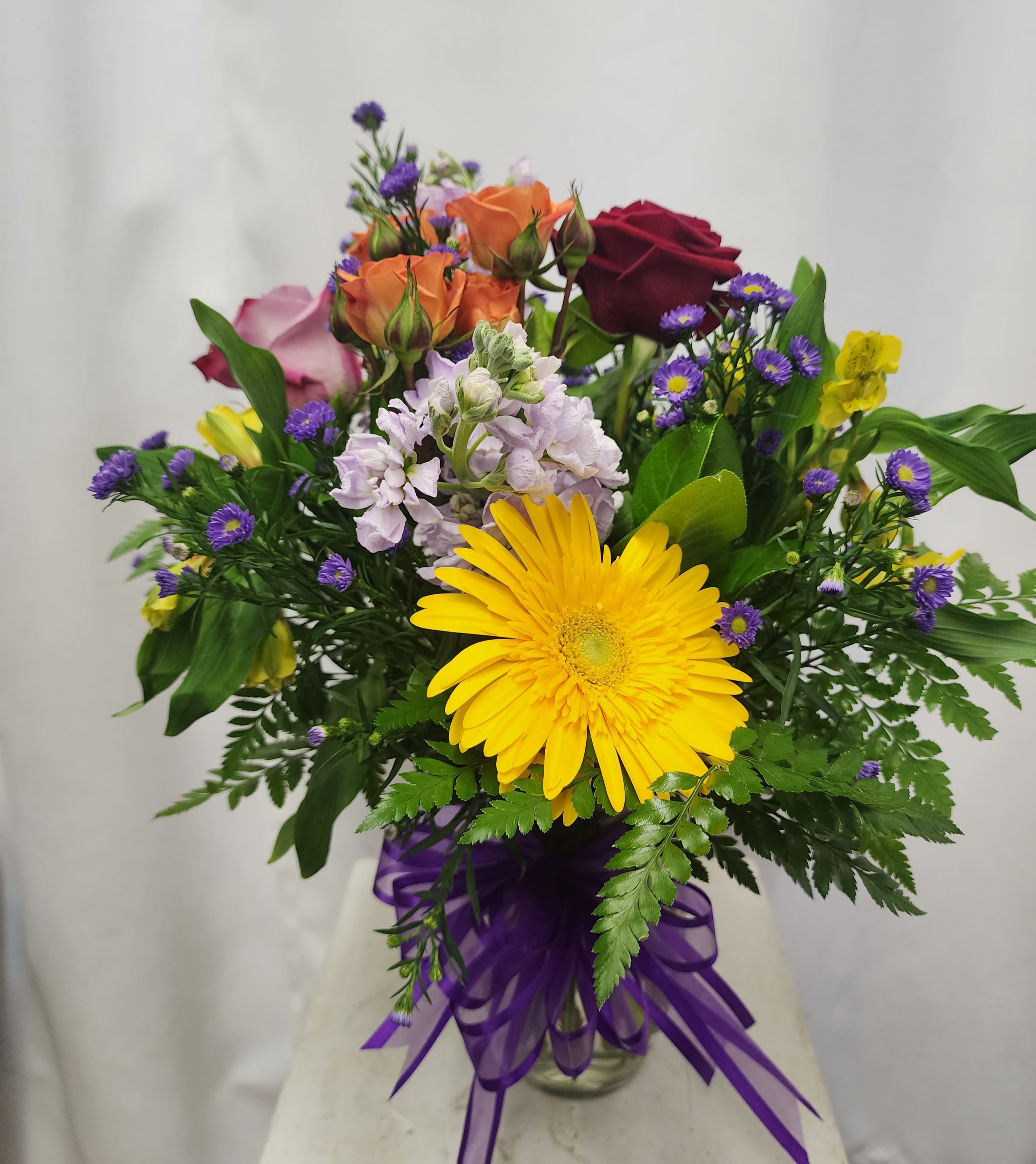 Mixed bouquet with a yellow gerbera daisy, roses, and purple filler flowers in a glass vase