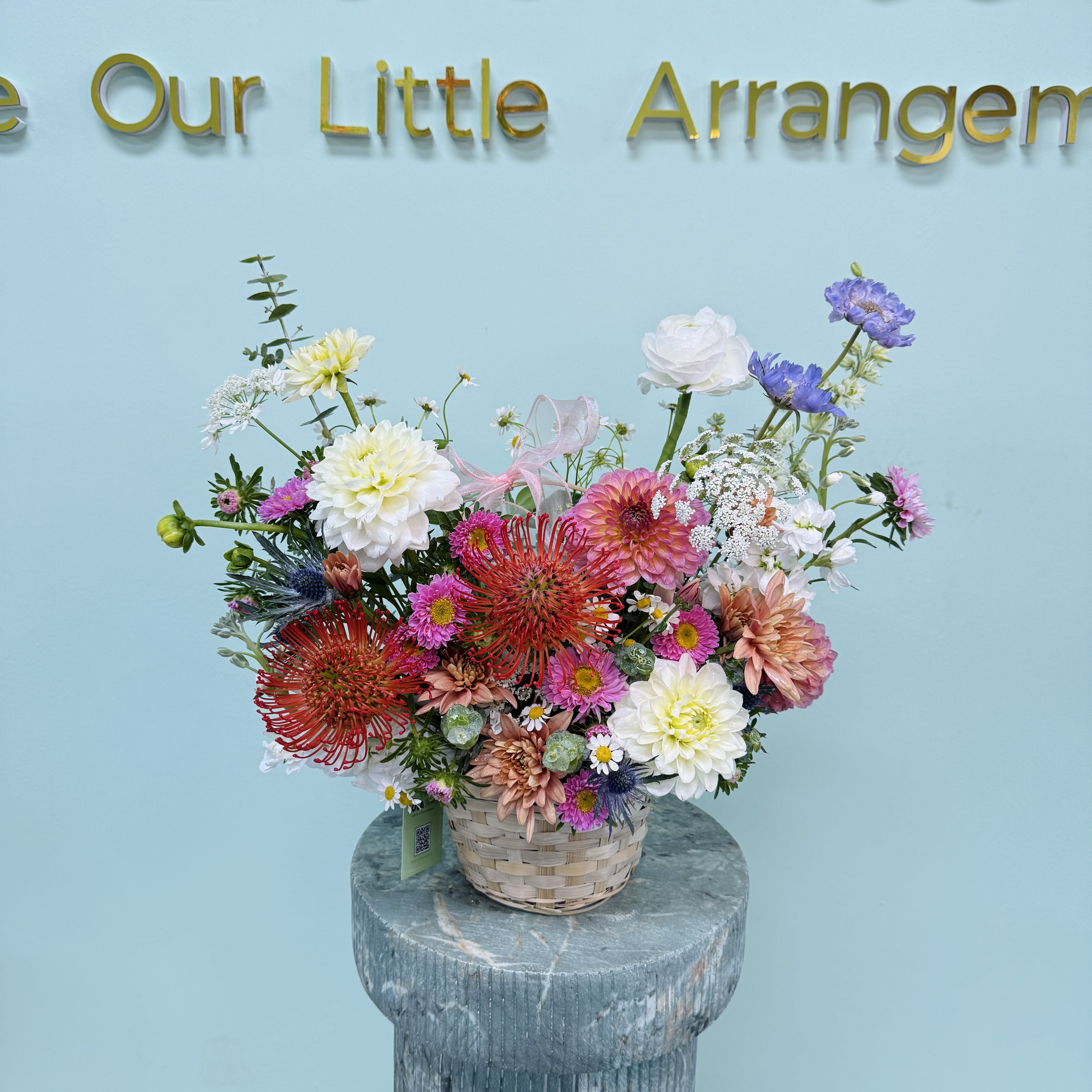 Custom Garden Style  - A lively, garden-style flower arrangement arranged low in a woven basket that instantly reads as handcrafted and heartfelt - perfect as a birthday bouquet, a thank-you gift, or a just-because surprise. The mixed-color palette of white, pink, purple, peach and touches of red creates an immediate cheerful impression; the recipient will feel genuinely celebrated the moment they see the layered, abundant display.  This piece mixes large chrysanthemum-like blooms with small daisy accents and spiky, textural focal flowers to give depth and movement. Presented in a sturdy woven basket with a soft pink ribbon tucked near the center and a gift tag, the arrangement feels casual and thoughtfully composed - like flowers gathered straight from a seasonal garden. The compact, low profile makes it an easy centerpiece for a table or a bright addition to a bedside or kitchen counter.  Ideal for spring occasions or whenever you want to say "I'm thinking of you," "thank you," or "happy birthday" without being formal. Because it's arranged for immediate display in water-holding floral foam inside the basket, no vase is needed; refresh with a little water at the foam if the flowers look thirsty and keep out of direct heat to extend their life.