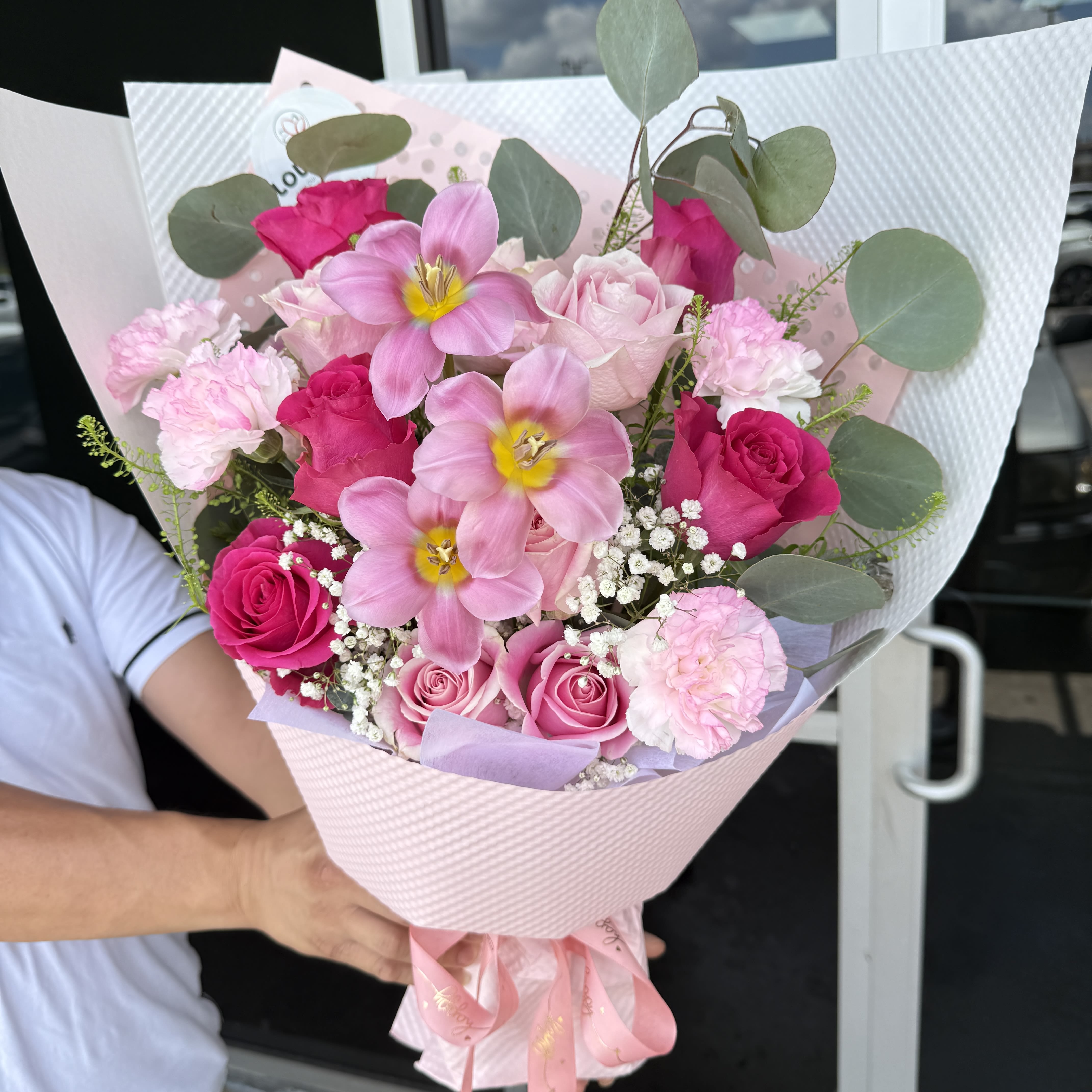 Bouquet 011 - Bright pink roses and soft pink tulip-like blooms make this birthday bouquet feel immediately joyful and romantic-perfect as an anniversary, Valentine's Day, or Mother's Day gift that says "you're cherished." The rounded, hand-tied design presents a feminine, polished look so the person you're thinking of will feel truly celebrated the moment they see it.  Pale pink carnations and clusters of white baby's breath add texture and a soft, cloud-like contrast to the richer roses, while eucalyptus-like greenery gives a fresh, herbal lift to the palette of pinks and whites. The stems are layered in decorative pink wrapping and finished with a ribbon for an elegant, ready-to-give presentation that looks beautiful from every angle.  Ideal for a mother, partner, or close friend who enjoys classic romantic blooms, this arrangement arrives ready to display-no arranging required. To keep it looking fresh, place the bouquet in clean water and trim stems every few days; available in a single hand-tied size as shown.
