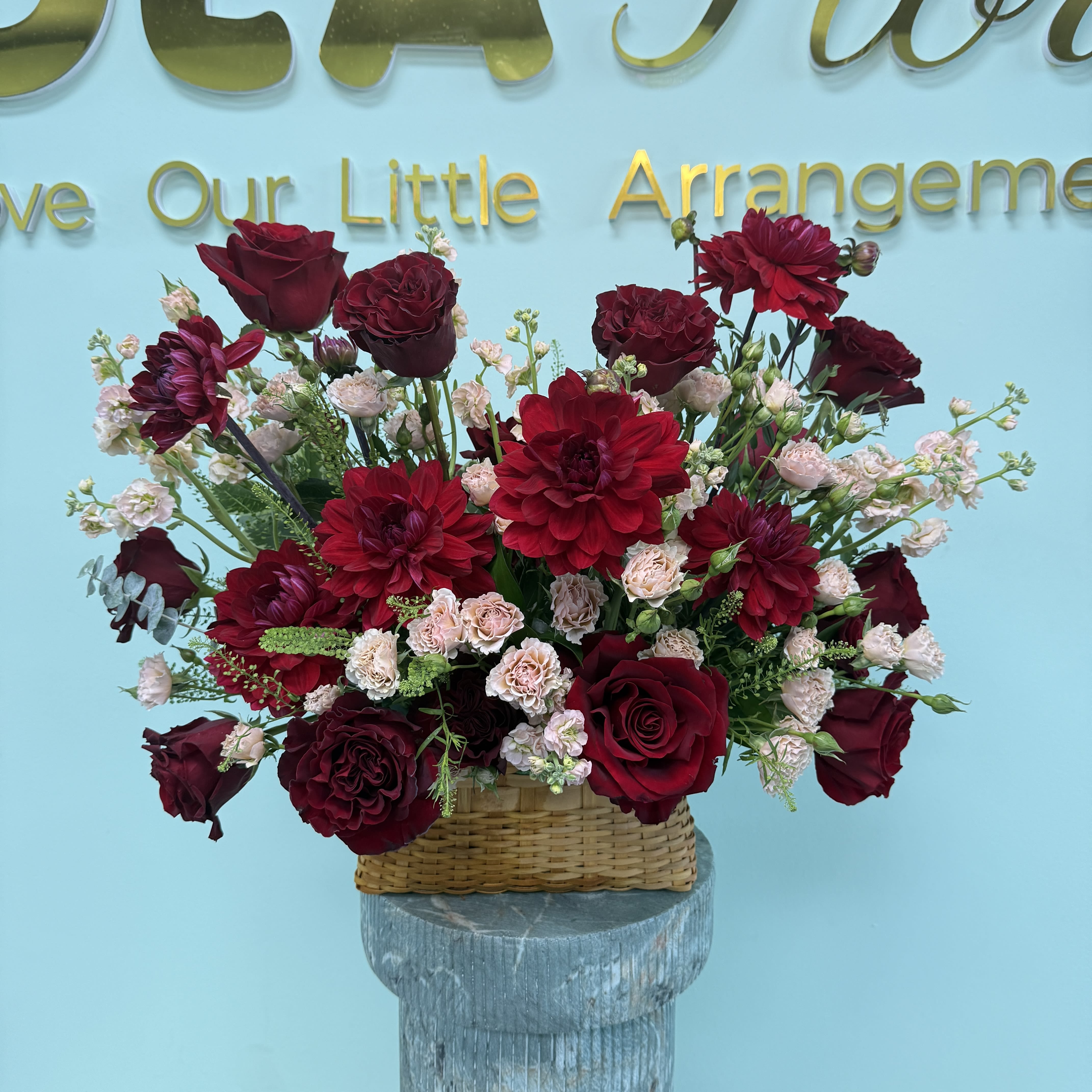 Red Roses Basket Bundles  - Deep, romantic red roses and velvety burgundy dahlias fill a low woven basket, creating a lush, rounded flower arrangement perfect for a birthday bouquet, anniversary roses, or a sympathy gift. The rich red-and-burgundy palette makes an immediate visual statement-intense and elegant-while the pale pink and white filler blooms soften the composition so the recipient feels both seen and comforted.  This basket arrangement pairs classic roses with textural dahlias and touches of carnations and delicate filler flowers in pinks and whites, set against fresh green foliage for contrast. The low, wide silhouette sits neatly on a table or mantle and reads as both decorative and emotionally thoughtful; sizing is compact but dense, designed to look full from every angle.  Send this as a romantic gesture, a meaningful way to say you're thinking of someone, or to mark a solemn occasion with care. The woven basket is ready to display-simple water maintenance will keep blooms fresh-so the person you choose will enjoy a polished, long-lasting arrangement without fuss.