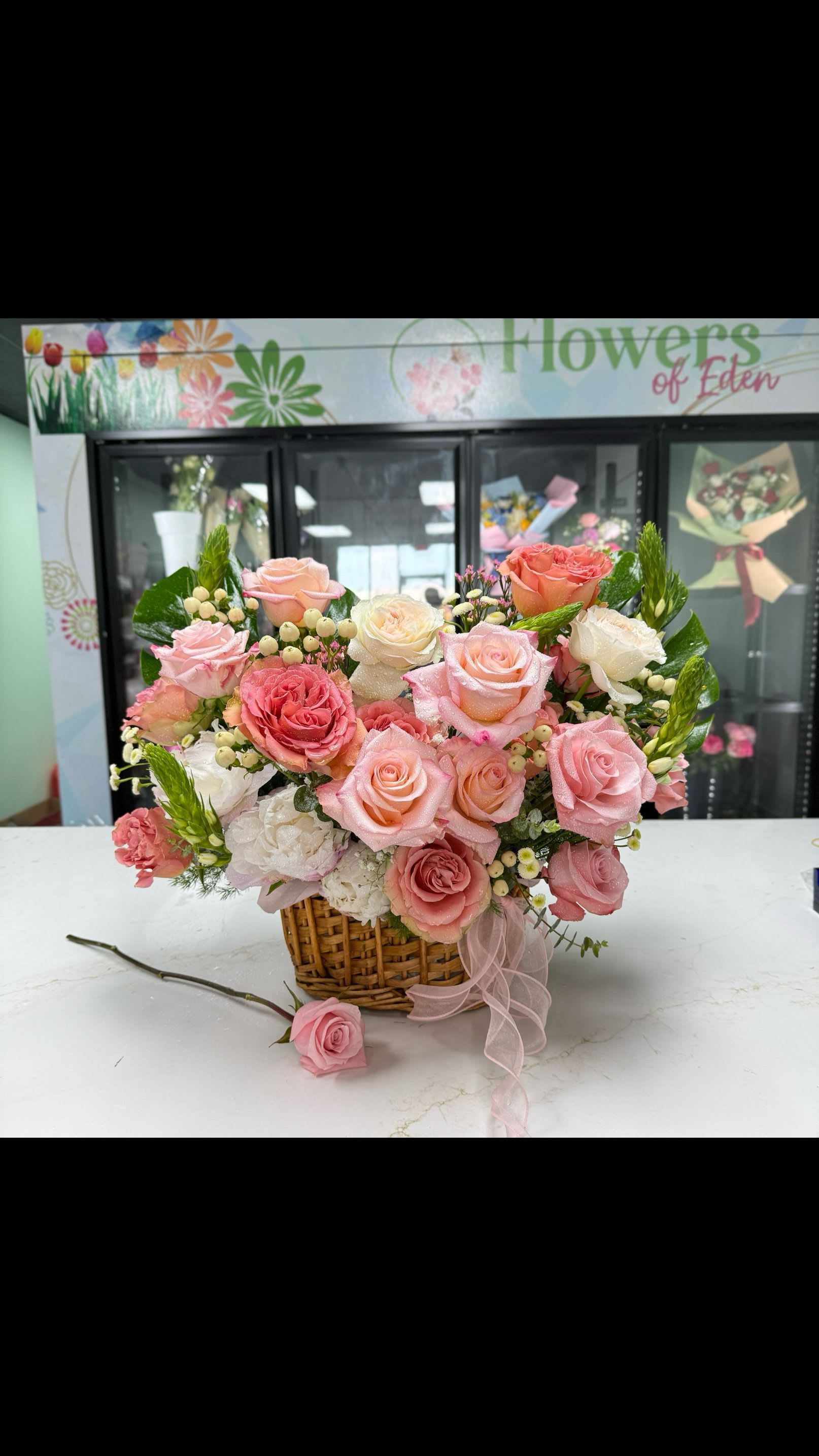 Roses Basket 03 - A lush cluster of pink, blush, and white roses-softly punctuated by ruffled carnations and tiny berry-like fillers-creates a romantic birthday bouquet or anniversary gift that feels both handcrafted and polished. The rounded, abundant shape in gentle pastels reads as affectionate and celebratory, making it the kind of arrangement that will make the person you're thinking of feel truly seen and cherished.  Arranged densely in a woven wicker basket and finished with a soft pink ribbon drape, this floral gift blends classic texture with a feminine, cottage-style charm. The mix of roses and carnations gives you visual depth and a variety of petal textures, while the small berries add delicate contrast against the pale palette of pinks, blush, white, and green foliage.  Perfect for birthdays, anniversaries, or a warm "just because" surprise, this cheerful everyday bouquet arrives ready to display in its basket-no vase required. To keep it fresh, change the water in the basket's liner daily, trim stems at an angle if you transfer to a vase, and keep away from direct heat; she'll enjoy the soft scent and full, long-lasting look for days.