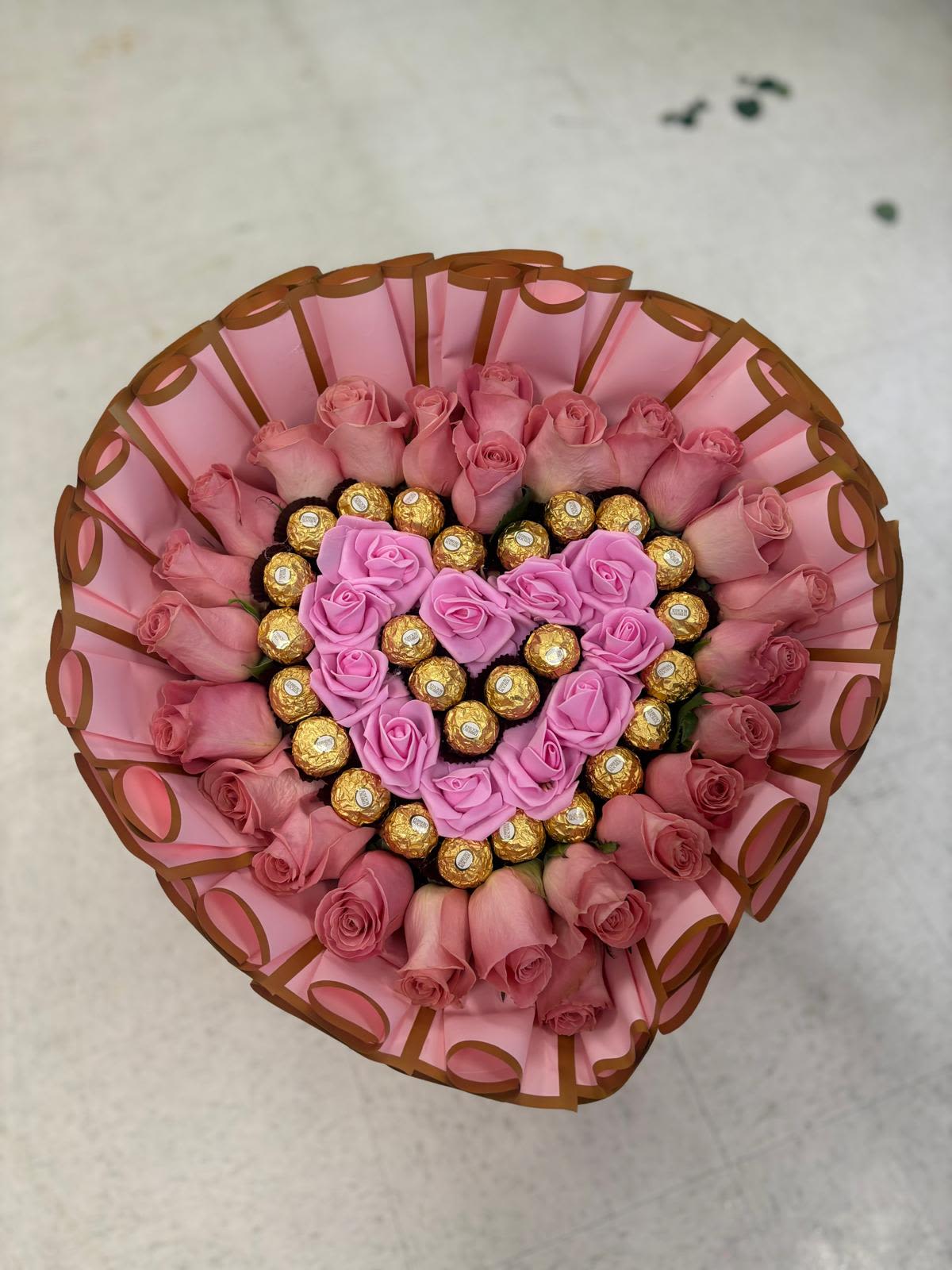 Chocolate heart and artificial flowers  - A heart-shaped rose arrangement romantic bouquet or a sweet surprise for anniversaries . The design centers on a lavender-pink heart of roses and gleaming gold-wrapped chocolates, surrounded by a fuller outer ring of soft pink . 
