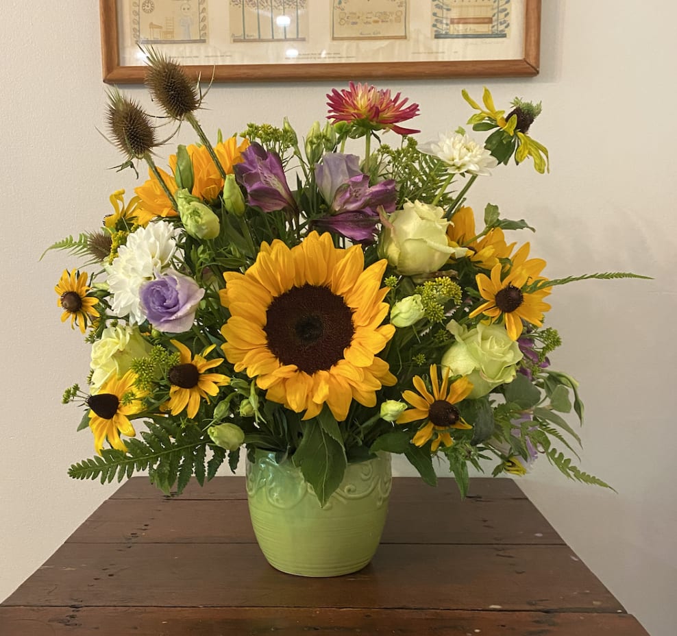 Mixed arrangement with sunflowers, yellow daisies, roses and purple blooms in a green ceramic vase