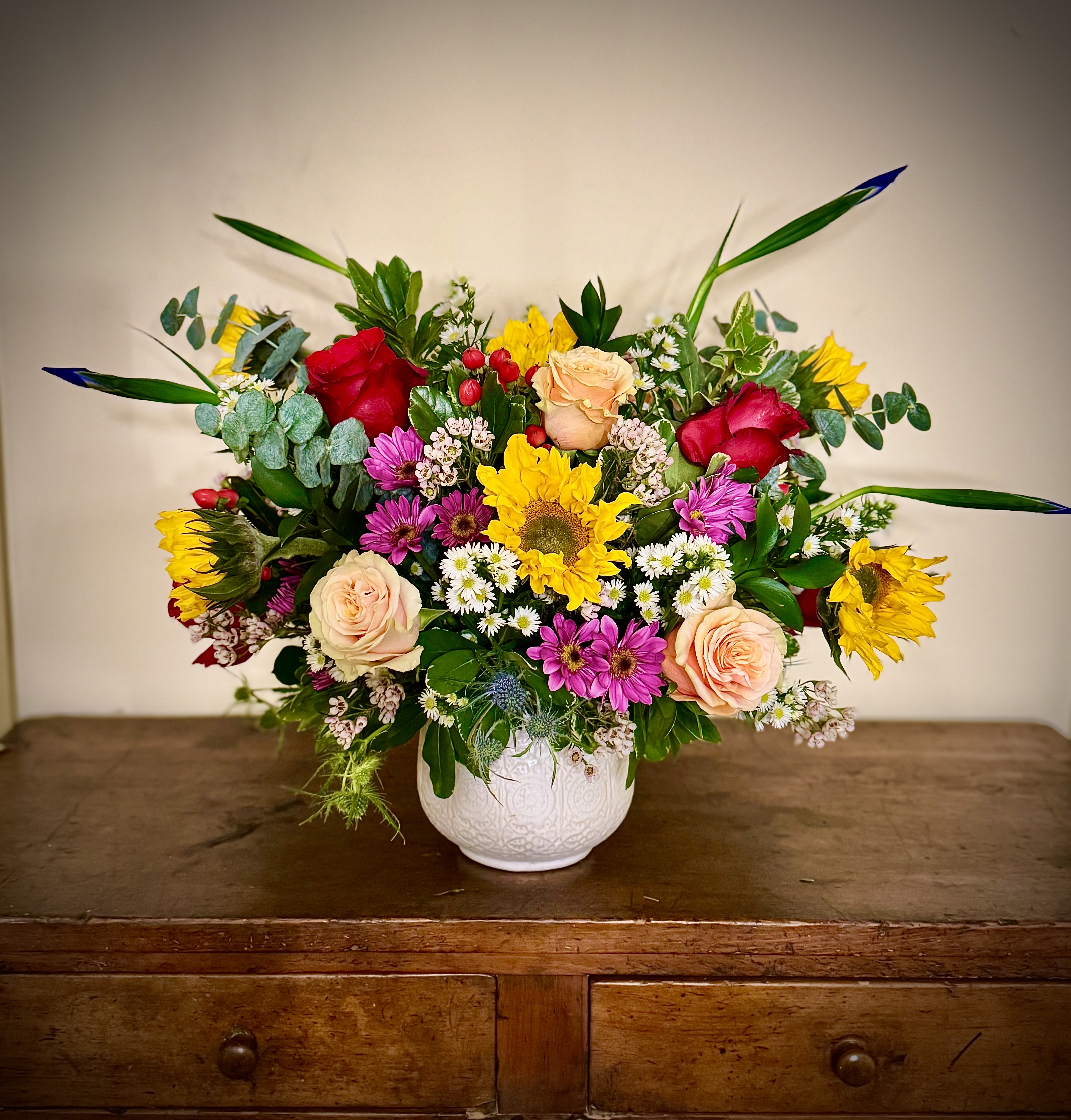 Colorful mixed arrangement with sunflowers, roses, and daisies in a white ceramic vase on a wooden table.