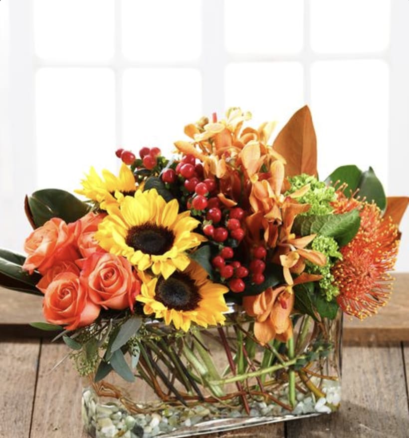 Low rectangular arrangement of sunflowers, orange roses, orchids, and berries in a clear glass vase.