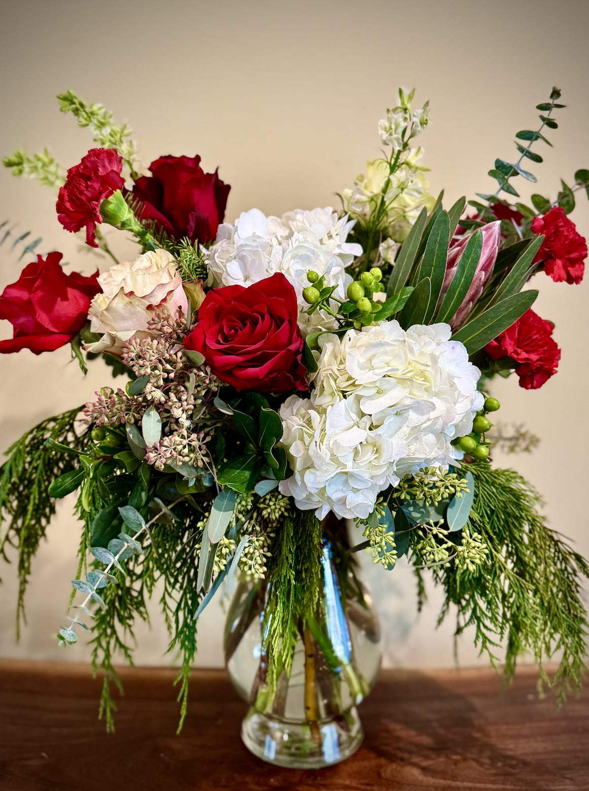 Tall arrangement of red roses, red carnations, white hydrangeas and evergreen foliage in a clear glass vase