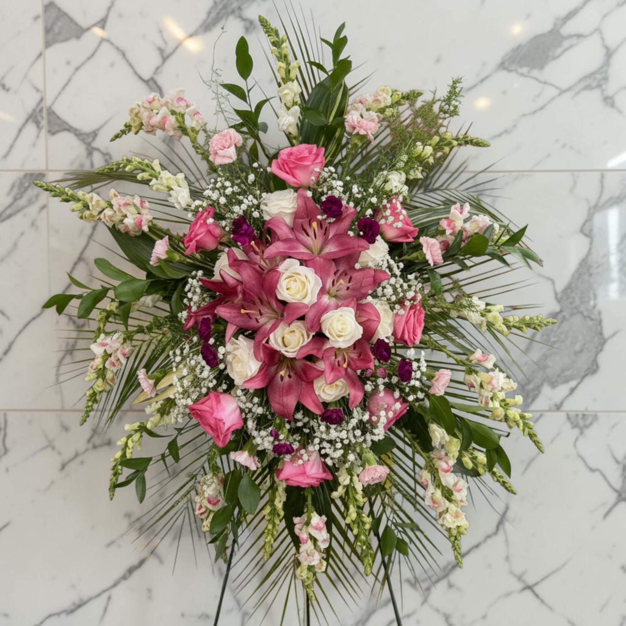 Compassion - Express your heartfelt condolences with this elegant standing spray, thoughtfully designed in soft shades of pink, white, and lavender. Featuring graceful lilies, roses, snapdragons, carnations, and delicate baby's breath, this beautiful tribute is accented with lush greenery for a timeless and comforting display. Arranged on a standing easel, it offers a meaningful expression of love, remembrance, and sympathy.
