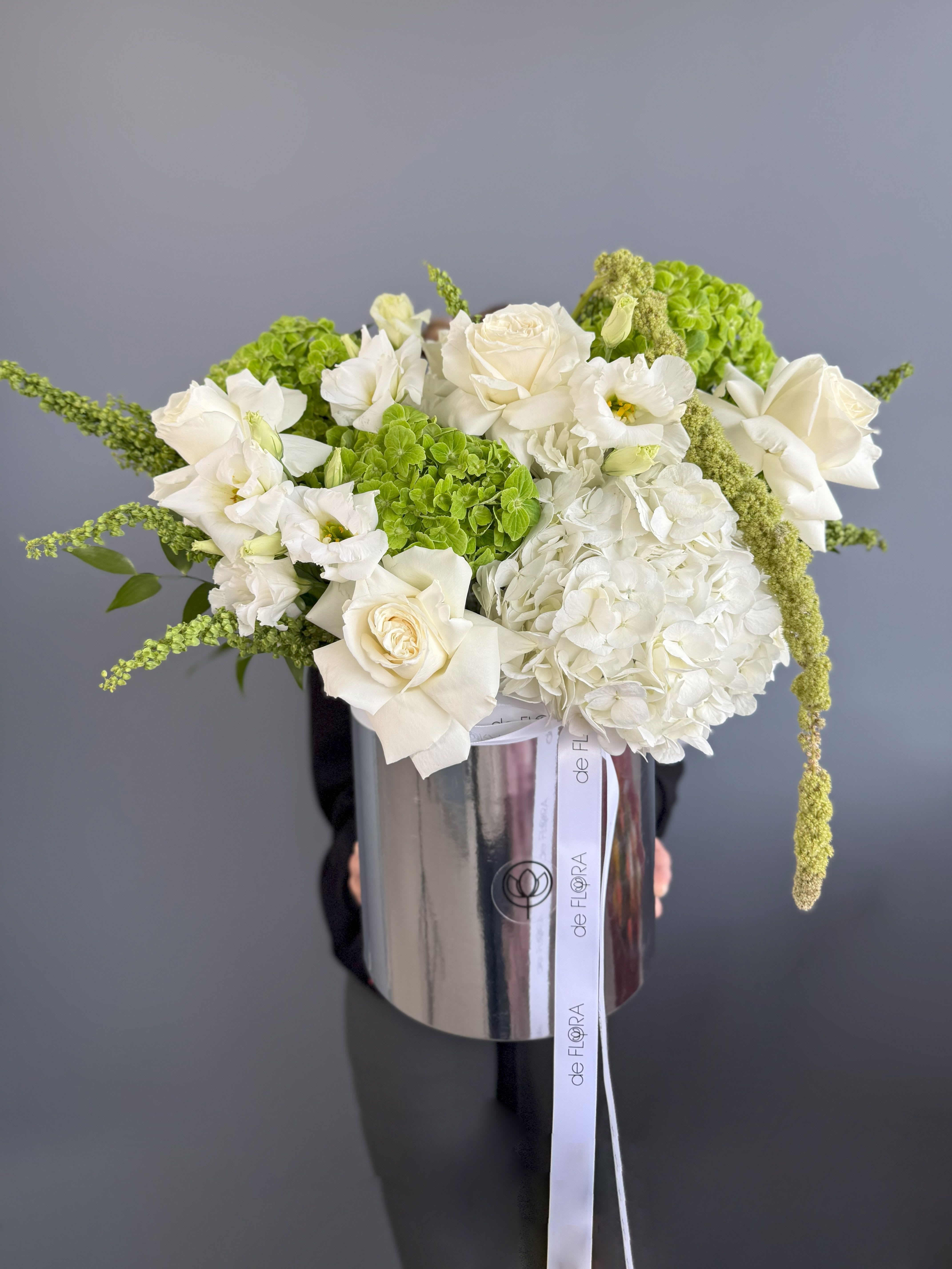 White and green flower arrangement with roses and hydrangeas in a tall silver hatbox-style container