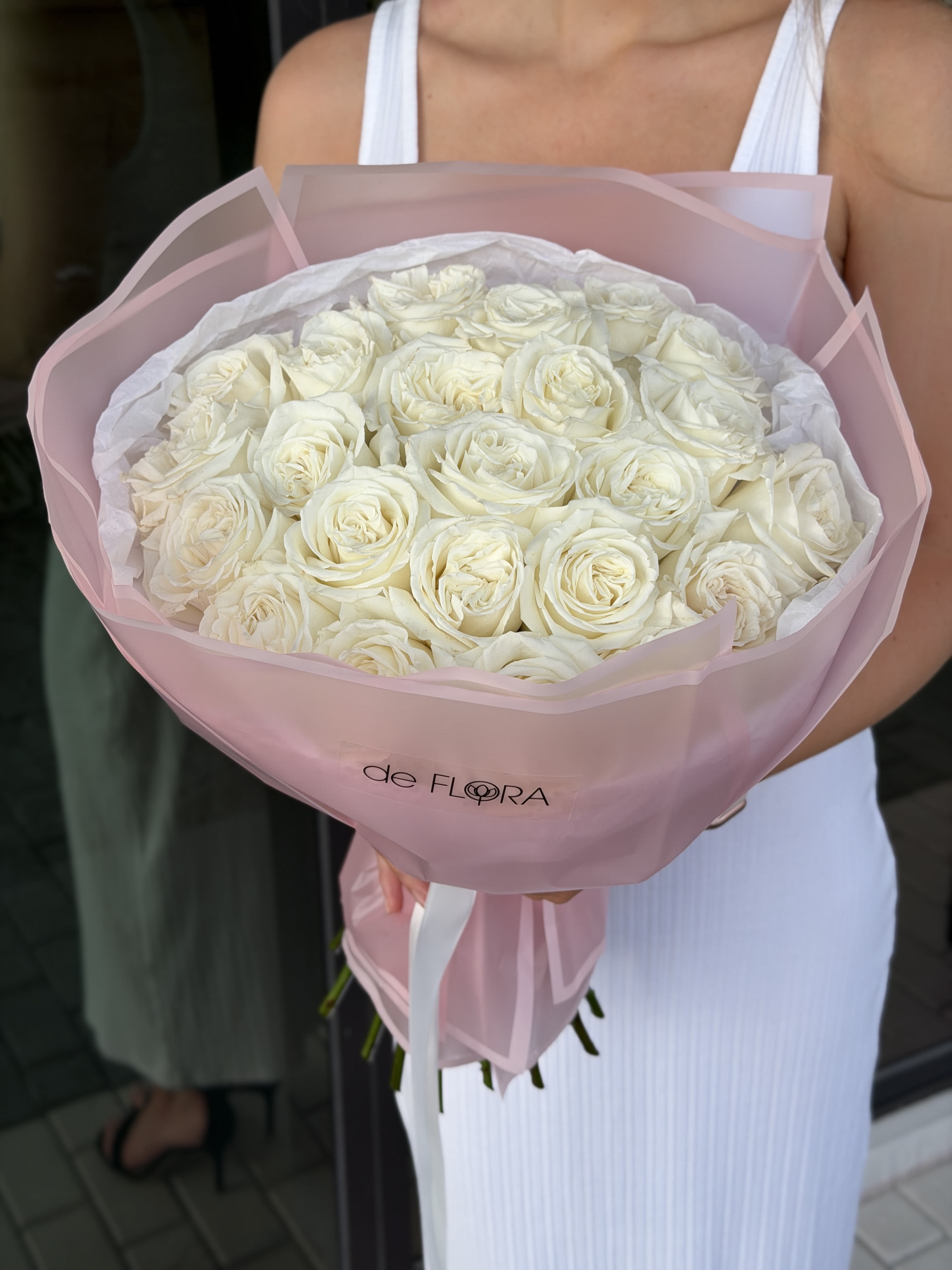 Large round bouquet of white roses wrapped in pale pink paper, held by a person in a white dress