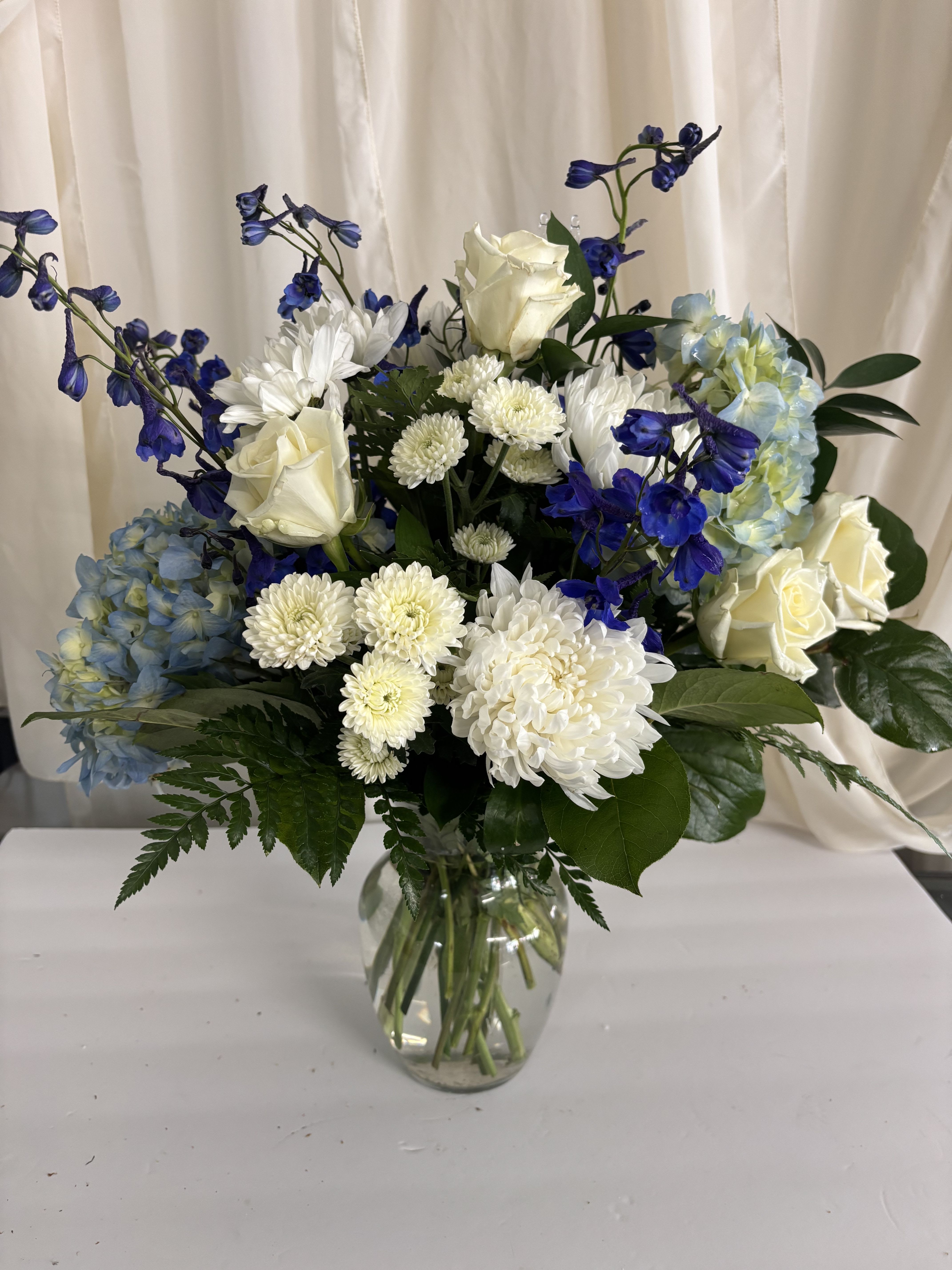 Cold as Ice - A clear glass vase that showcases clustered white roses, chrysanthemums, pale blue hydrangea, and deep blue accents for a fresher, more formal look.