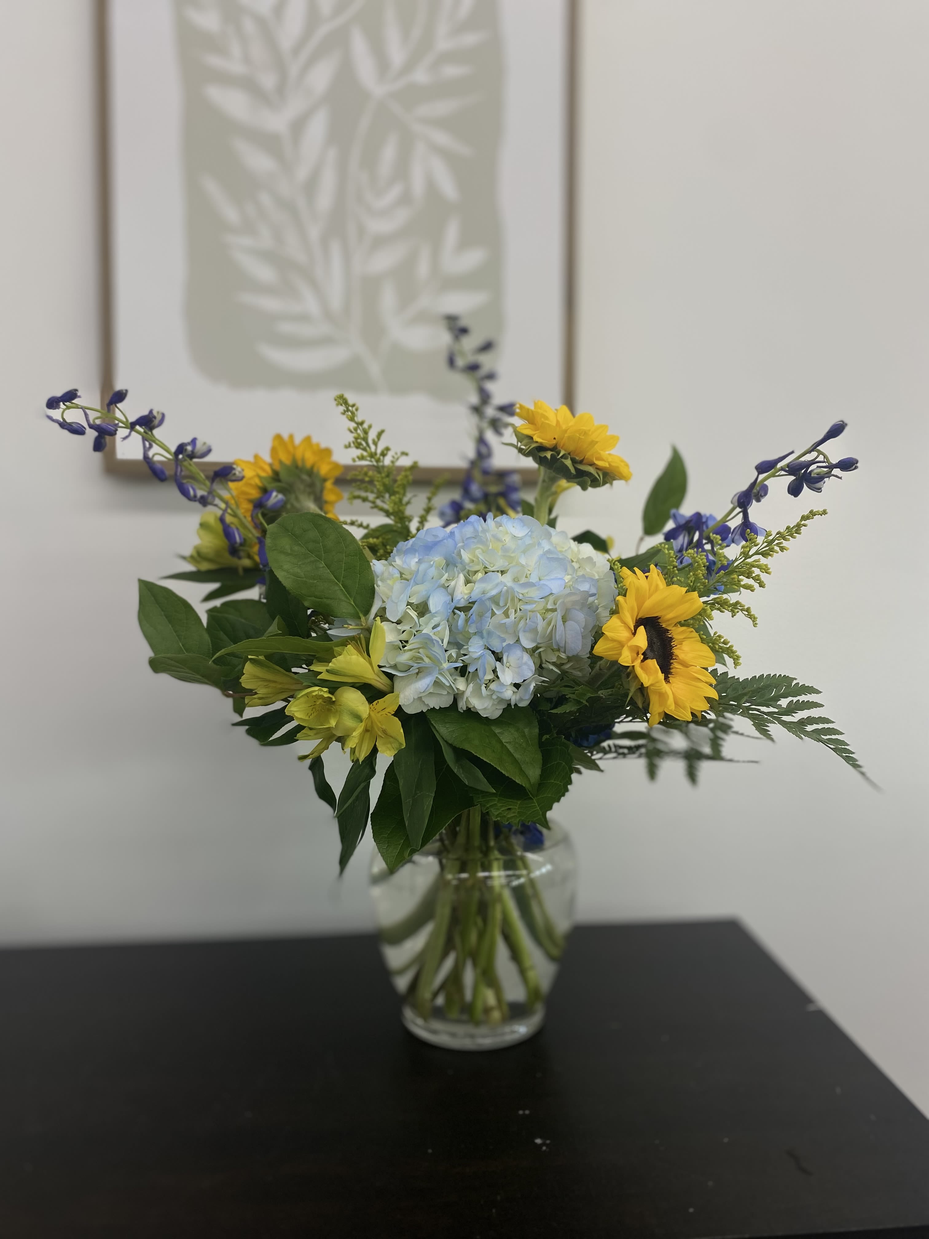 Here comes the "Son" - Welcome home a sweet baby boy with this bright and sunny arrangement. Filled with a mix of blue flowers accented with gorgeous sunflowers, this piece is sure to make the new little one feel right at home. 