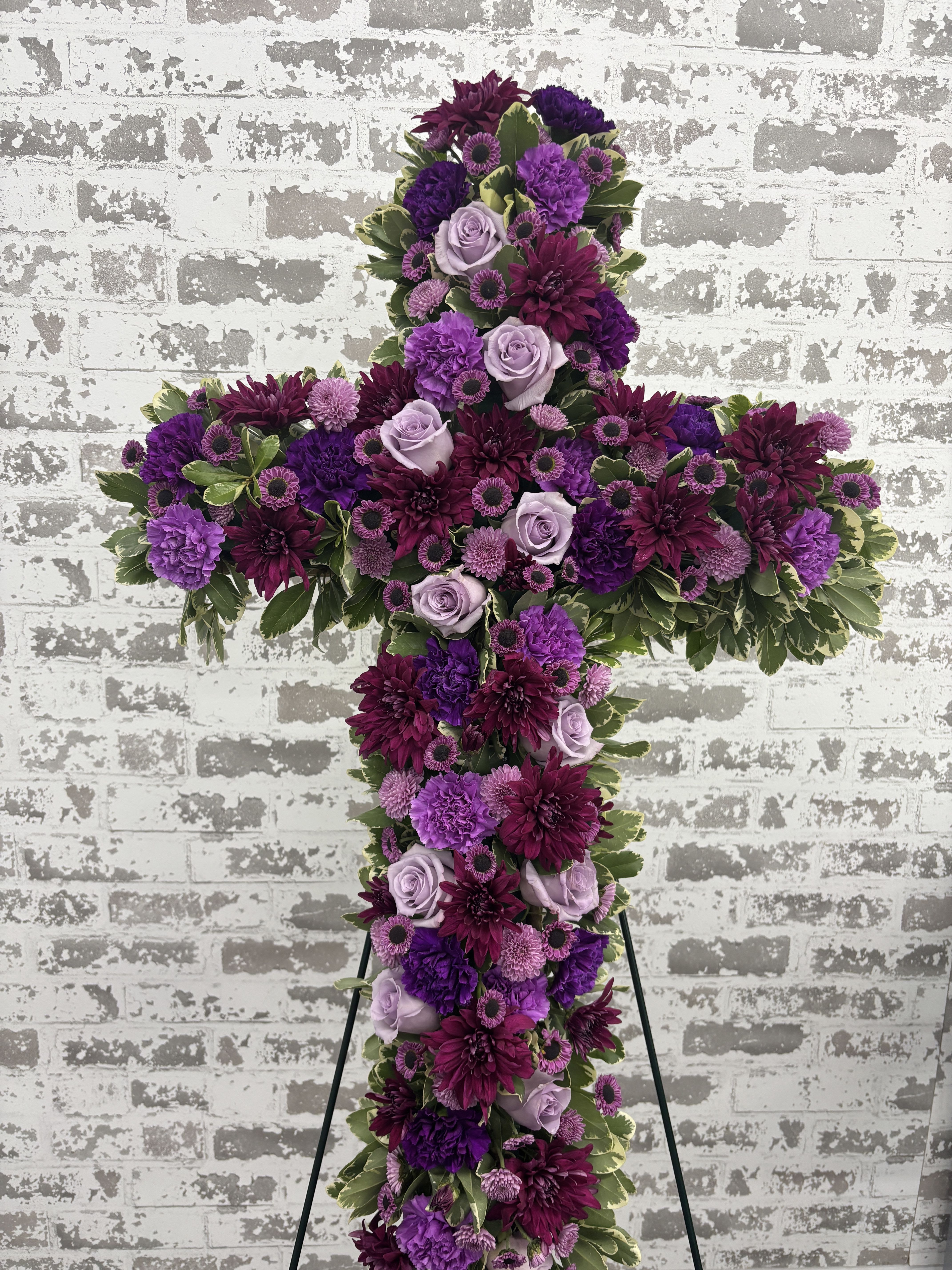 Purple &amp; Lavender Cross Sympathy Floral Arrangement - This striking tribute features a beautiful blend of premium fresh flowers, including lavender roses, carnations, chrysanthemums, button poms, and complementary seasonal blooms, accented with lush, variegated greenery. The cross is approximately 30–36 inches. It includes around 10–15 roses and a generous number of premium flowers. The easel is included.  