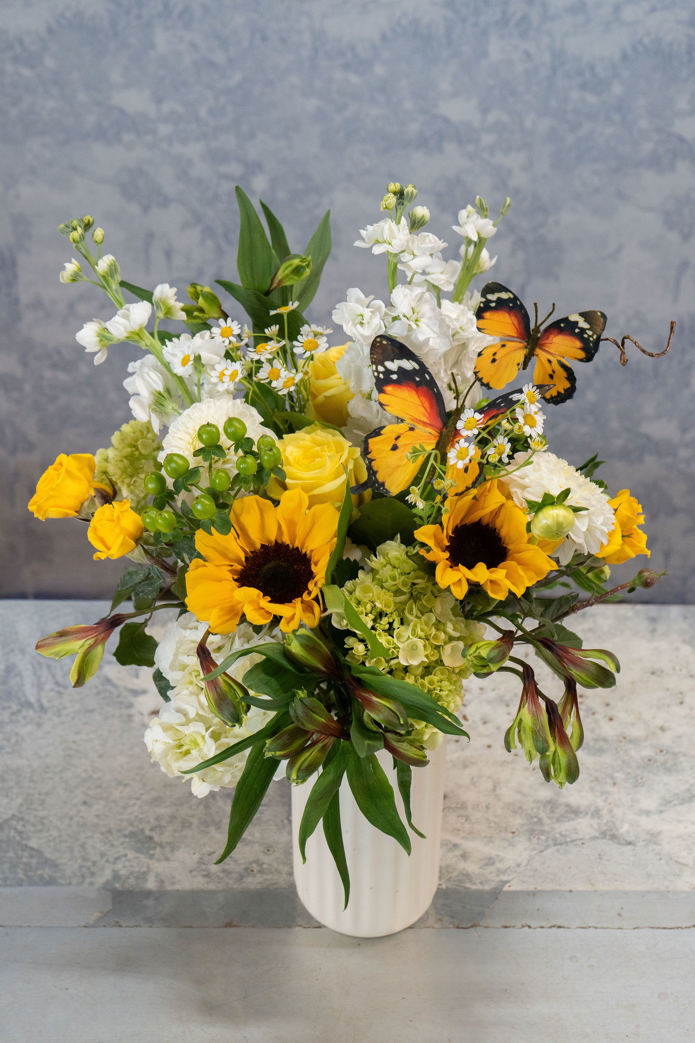 Butterfly Kisses - Sunflowers steal the show in this sunny flower arrangement, a cheerful birthday or thank-you gift that feels like a warm embrace. Designed for Mother's Day and everyday celebrations alike, the bouquet combines bright yellow sunflowers with soft yellow roses, crisp white blooms, and playful daisy-like accents to create an immediately uplifting impression your recipient will notice. Layered clusters of pale green hydrangea, rounded white chrysanthemums, and airy alstroemeria add texture and movement, while decorative butterfly picks lend a whimsical, garden-fresh touch. Arranged in a clean white ceramic vase, the compact composition balances bold color against dark sunflower centers for high visual impact without taking up much space.