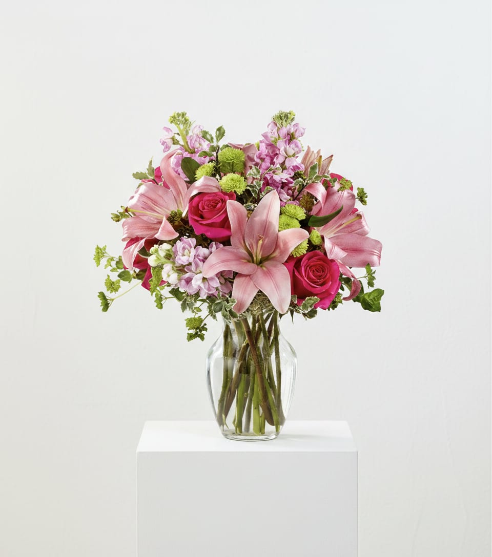 LUXE BLOSSOM  - Pink lilies and vivid pink roses take center stage in this Luxe Blossom birthday bouquet, creating a romantic, springlike surprise for anniversaries or to say you're thinking of her. Soft lavender stock and small green chrysanthemums weave through the arrangement, giving it a fresh, celebratory lift that reads equally as heartfelt for a milestone or a just-because moment.  The flowers are gathered in a rounded, full composition set in a clear glass vase that shows the clean stems and keeps the look airy and modern. Colors range from warm pinks to cool lavenders with crisp white highlights, and the mix of lilies, roses, stock, alstroemerias or daisies offers varied textures - velvety petals, frilly stock blossoms, and button-like green mums - for visual depth and softness.  Designed to make the recipient feel genuinely celebrated, this arrangement is ideal for mothers, partners, friends, or coworkers who appreciate a feminine palette with a joyful spirit. The clear glass vase sits on a white pedestal for an extra polished presentation; enjoy fresh water changes and trim stems every few days to extend vase life.
