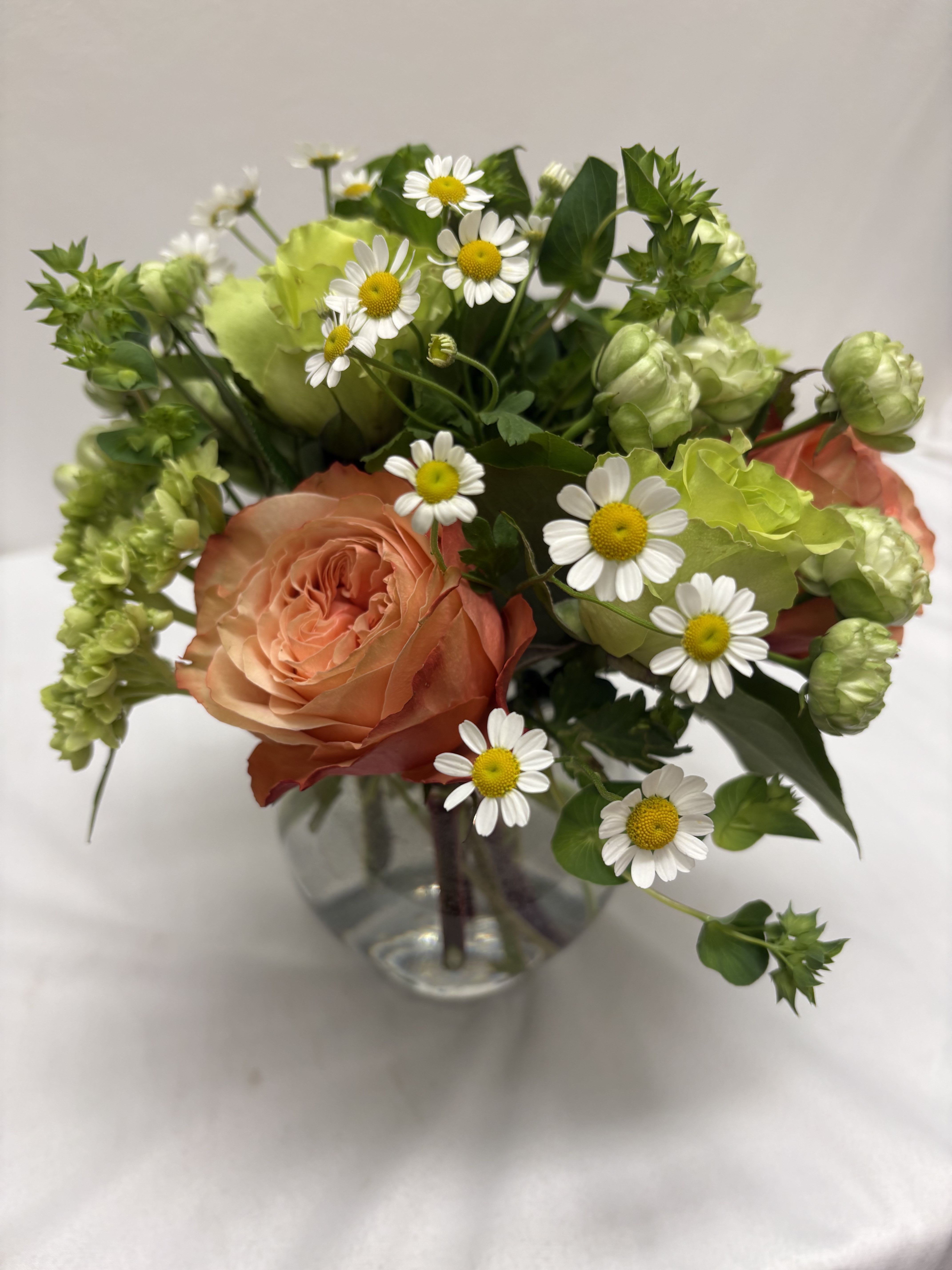 Small peach rose and white daisy arrangement with green accents in a round clear glass vase.