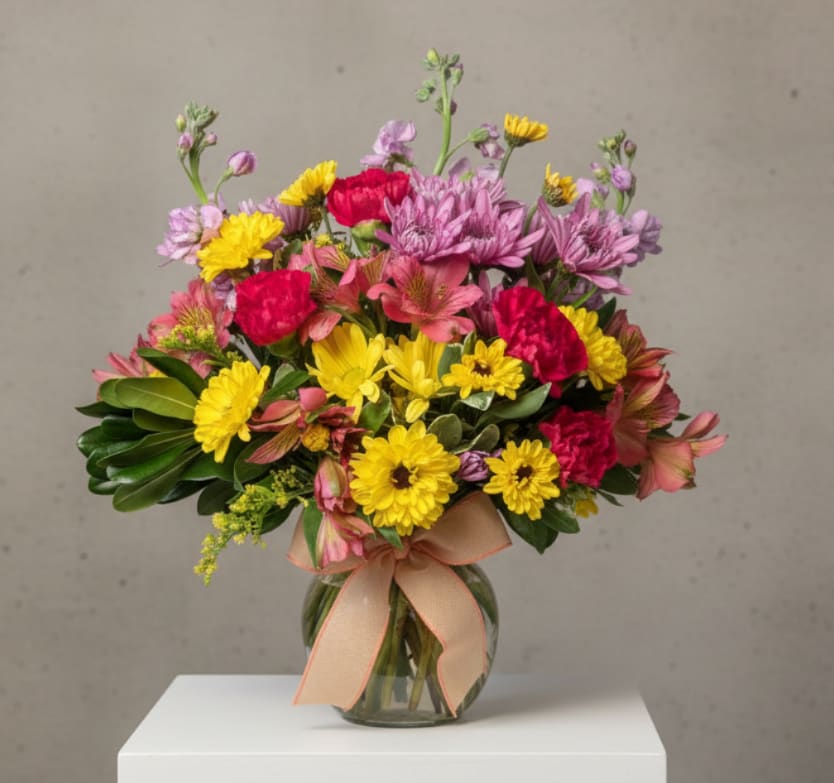 Mixed bouquet of pink, yellow, and purple flowers in a glass vase with a ribbon