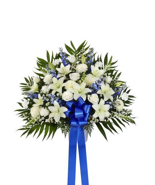 Standing spray of white lilies, roses, and carnations with blue flowers and a blue ribbon bow