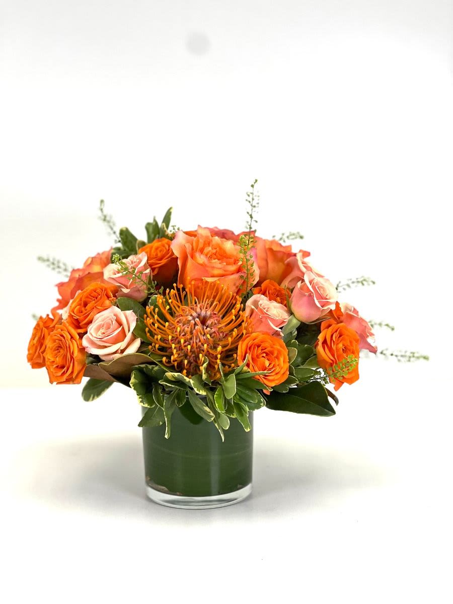 Low arrangement of orange and peach roses with a pincushion protea in a clear glass vase