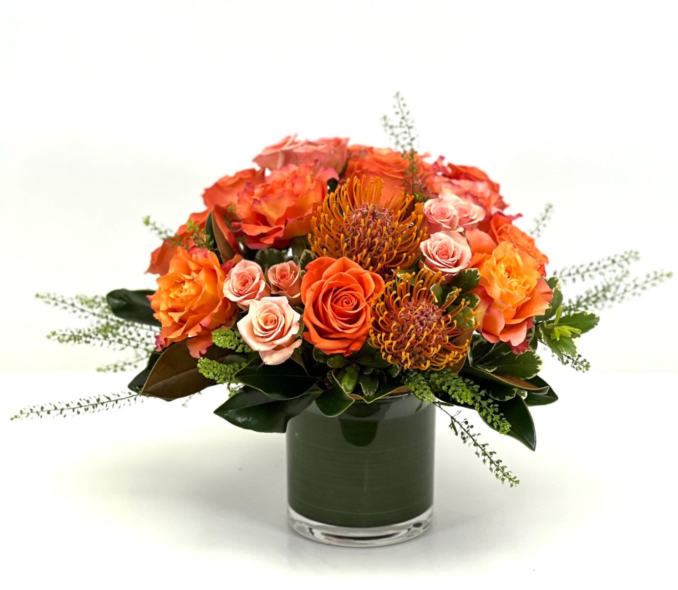 Low orange and peach rose arrangement with pincushion protea in a glass cylinder vase.