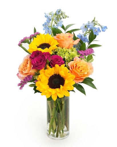 Tall glass vase of yellow sunflowers, peach and magenta roses, blue delphinium and mixed blooms