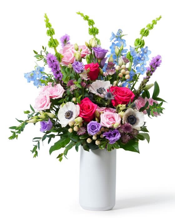 Tall mixed bouquet of pink roses, white anemones, and purple and blue blooms in a white vase
