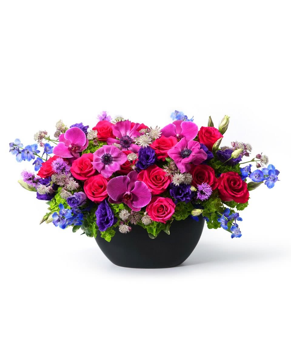 Low arrangement of hot pink roses, purple orchids, and blue flowers in a black bowl vase
