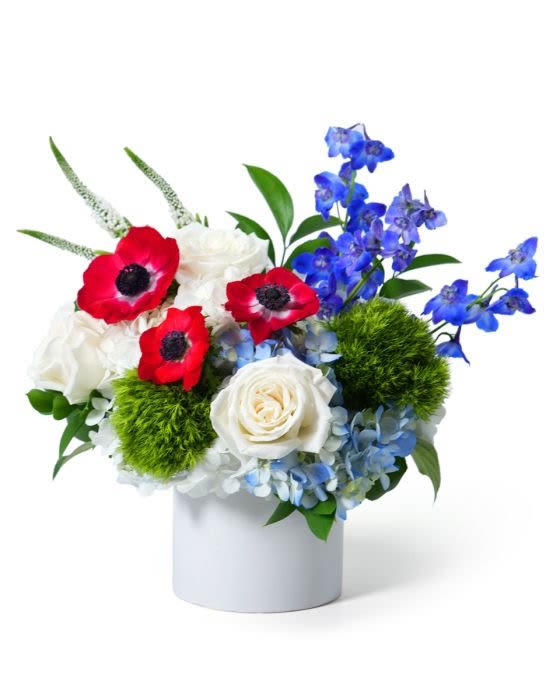 Low red, white, and blue flower arrangement with roses and anemones in a white cylinder vase