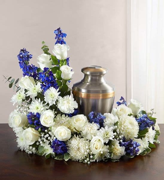 Cremation urn ringed by white roses, white mums, and blue flowers in a low sympathy arrangement.