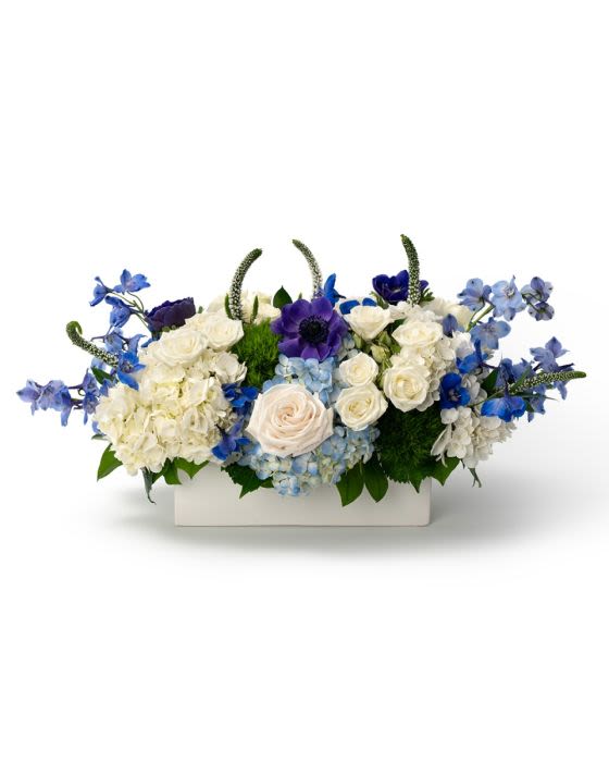 Low centerpiece of white and blue flowers, including roses and hydrangeas, in a rectangular white container