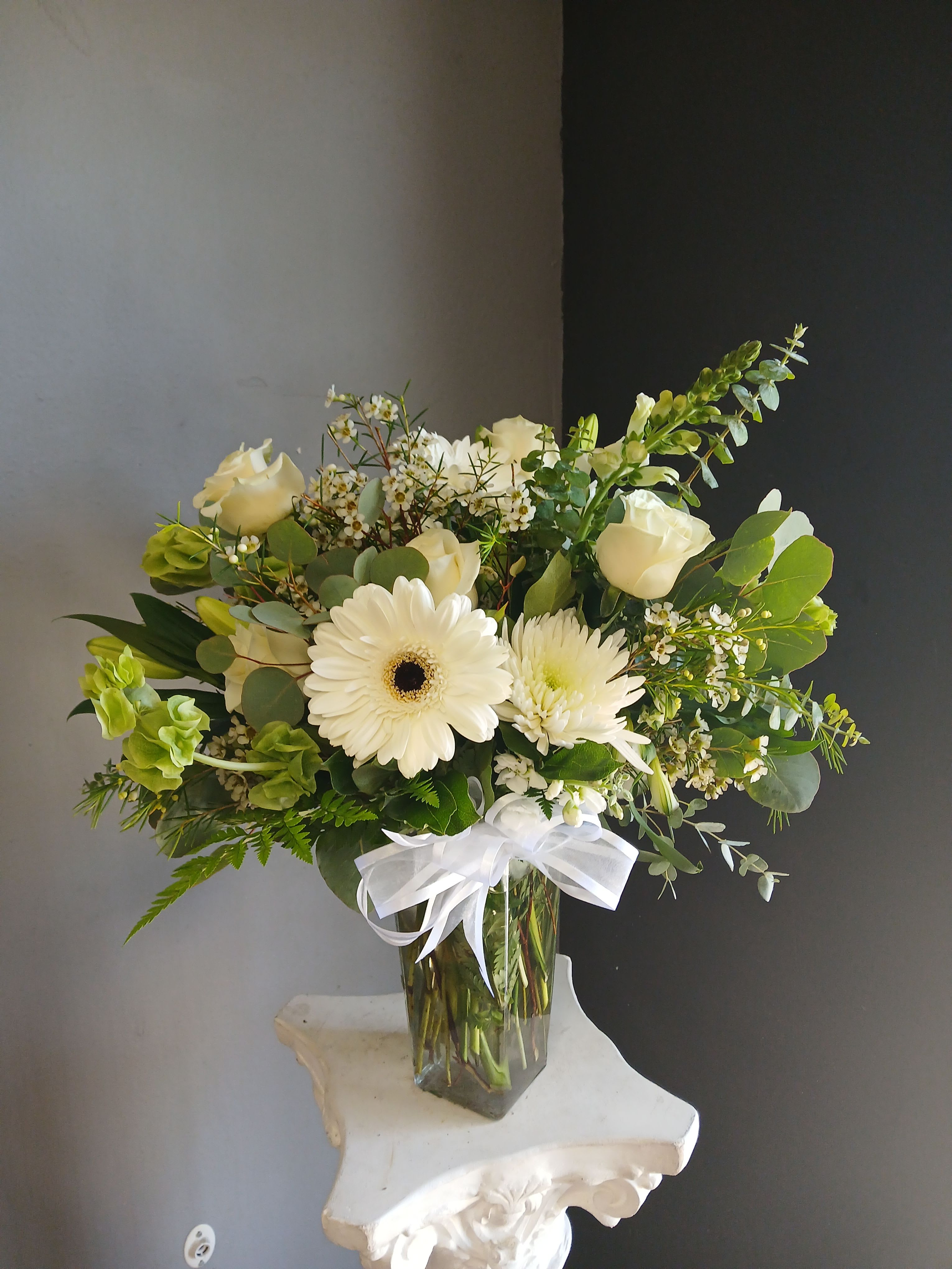 #799 All White Vase Arrangement  - Long trailing greens and pale yellow accents give movement against a clean backdrop of fresh foliage. The stems arrive arranged in a clear square glass vase that frames the airy silhouette and makes a simple, modern statement.  Choose this arrangement to offer quiet support, mark a subtle celebration, or brighten someone's home without overwhelming their space. It's presented ready to display in the included vase; for best longevity, keep water fresh and out of direct heat or sunlight to preserve the blooms' clean, soft look.