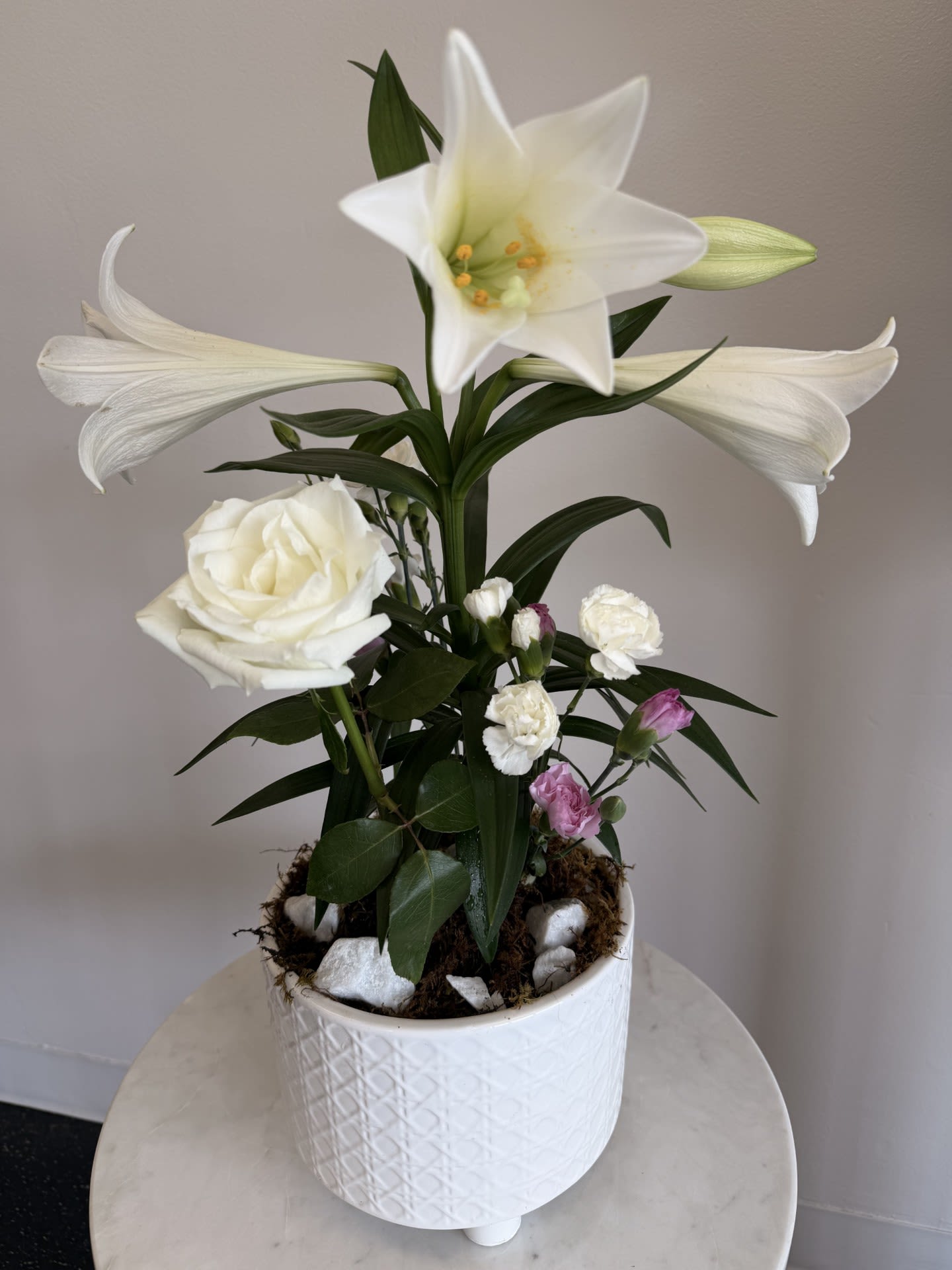 Easter Lily Plant  - The white Easter lily with it's beautiful trumpet-shaped blossoms symbolizes purity, hope, and life - the spiritual essence of Easter - and all the promises of Spring. Whether given as a gift or enjoyed in your own home, the Easter Lily also know as the Peace Lily serves as a beautiful reminder that Easter is a time for rejoicing and celebrating. One white Easter lily arrives in a decorative ceramic pot dressed up with fresh cut flowers, moss &amp; rocks to elevate its elegance even more!   WHILE SUPPLIES LAST 