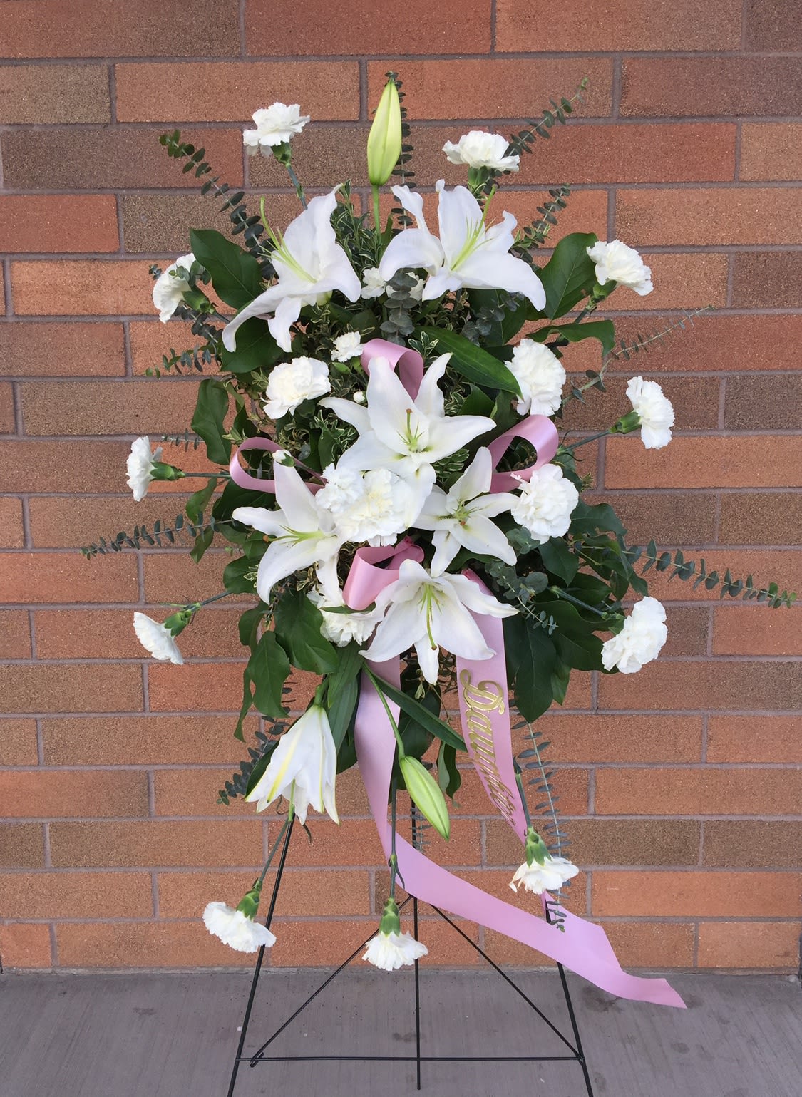 Standing white lily and carnation spray with pink ribbon on an easel