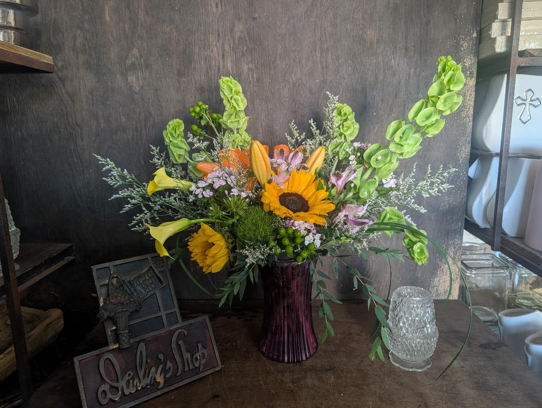 Sofia - Bright sunflowers and yellow calla lilies create an instant burst of sunshine in this everyday mixed bouquet, perfect for a birthday, thank-you, or general celebration. The arrangement's cheerful yellow and orange blooms-accented with soft pink flowers-announce joy at sight, while the tall purple glass vase adds a striking contrast that makes the recipient feel especially seen.  The design is airy and garden-style with an asymmetrical silhouette: long green stems, branching filler, and orange lily blooms give movement and texture, and the soft pink alstroemeria (and other delicate accents) soften the palette. Presented in a tall purple glass vase, it stands roughly as a statement centerpiece while remaining light and loose in feel.  Send this when you want someone to smile-whether marking a small victory or brightening an ordinary day. The vase arrives ready to display; keep flowers in fresh water, trim stems every few days, and place away from direct heat to extend their lively color and shape.
