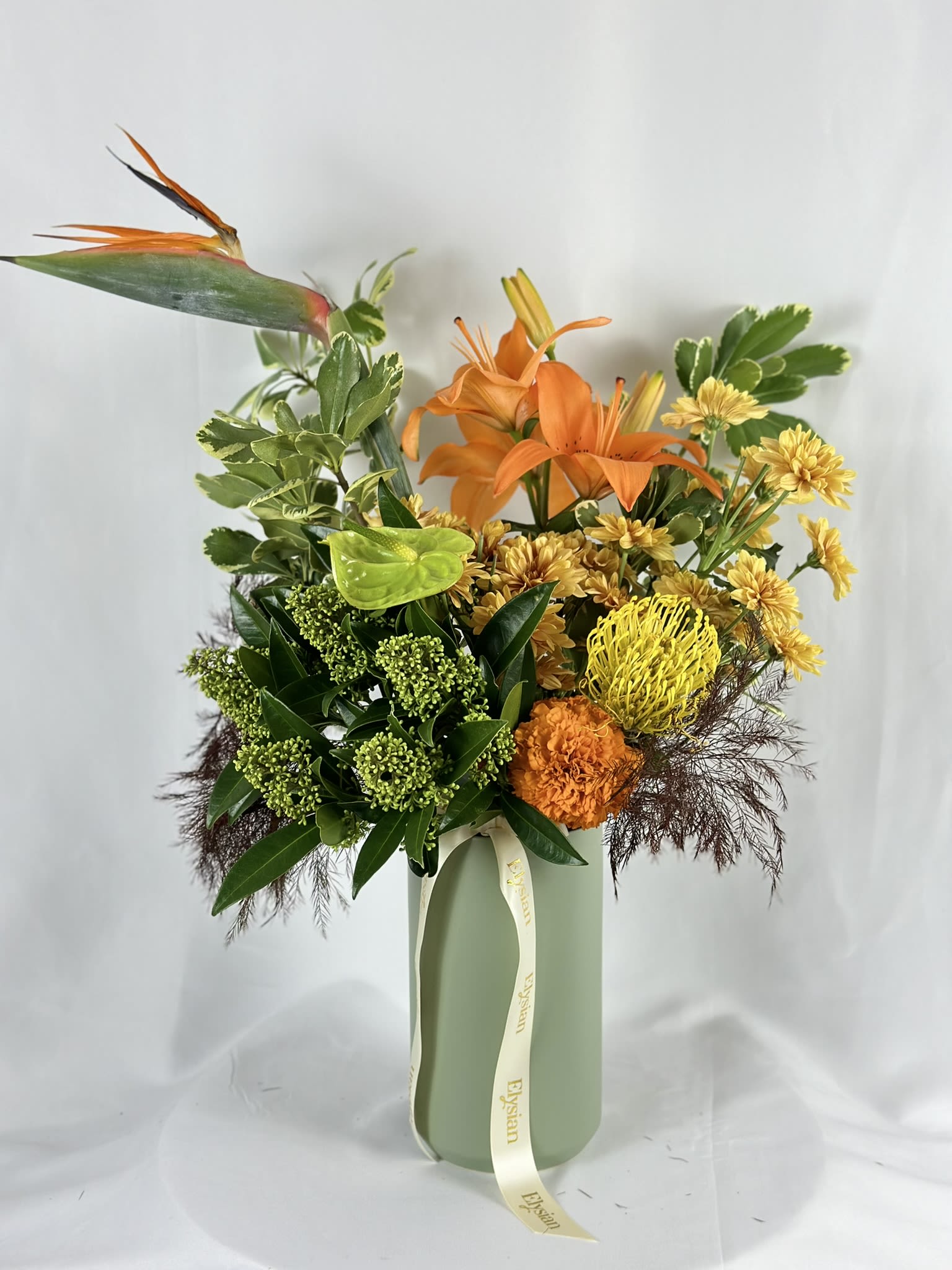 Tropicana - A single bird of paradise stretches skyward like a piece of living art, setting the tone for everything below: orange Asiatic lilies in full trumpet, golden cushion chrysanthemums, a vivid yellow pincushion protea, a lime green anthurium, deep orange carnations, skimmia buds, and a cascade of burgundy asparagus fern that grounds the whole composition in rich, earthy contrast.