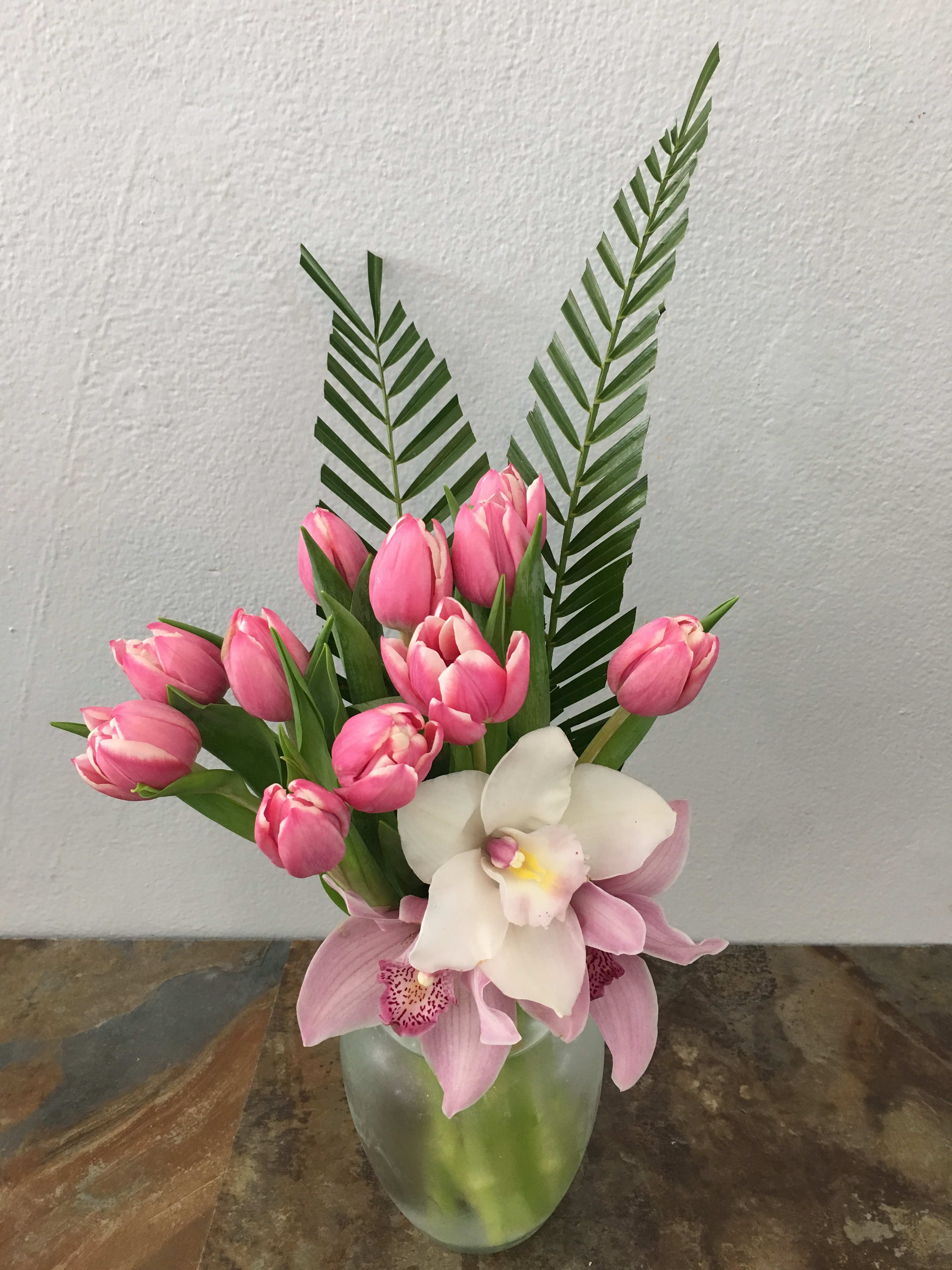 Pink tulips and orchids arranged in a glass vase with tall palm fronds.