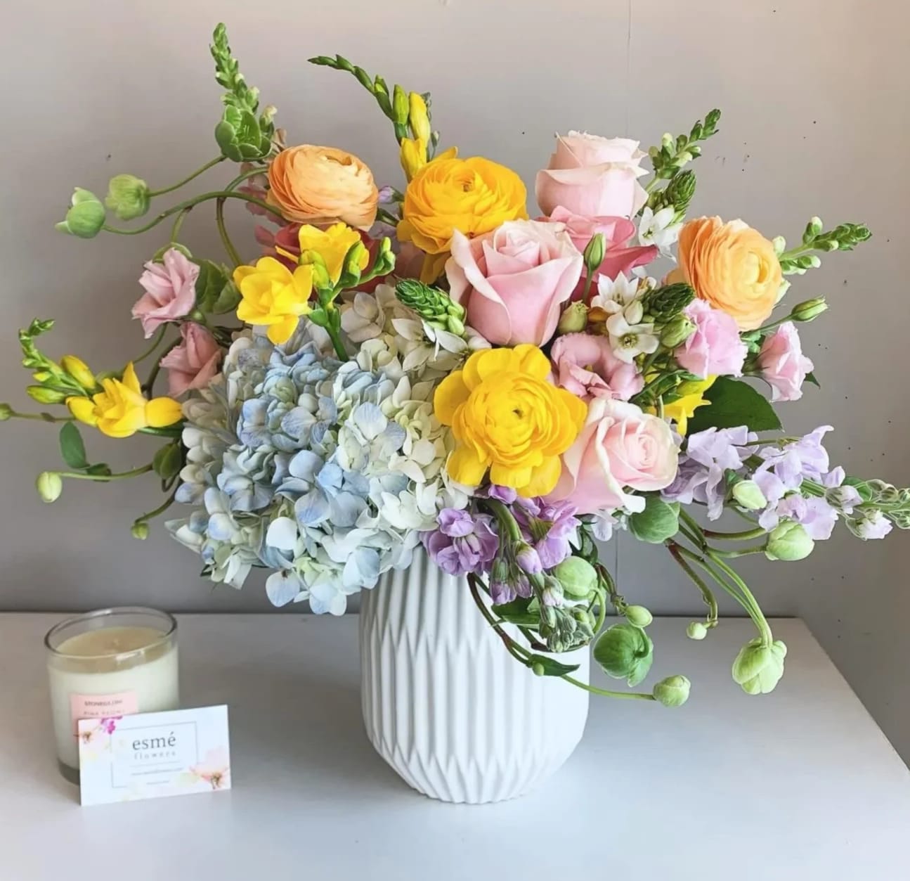 Mixed pastel bouquet in a white vase with a candle and card beside it