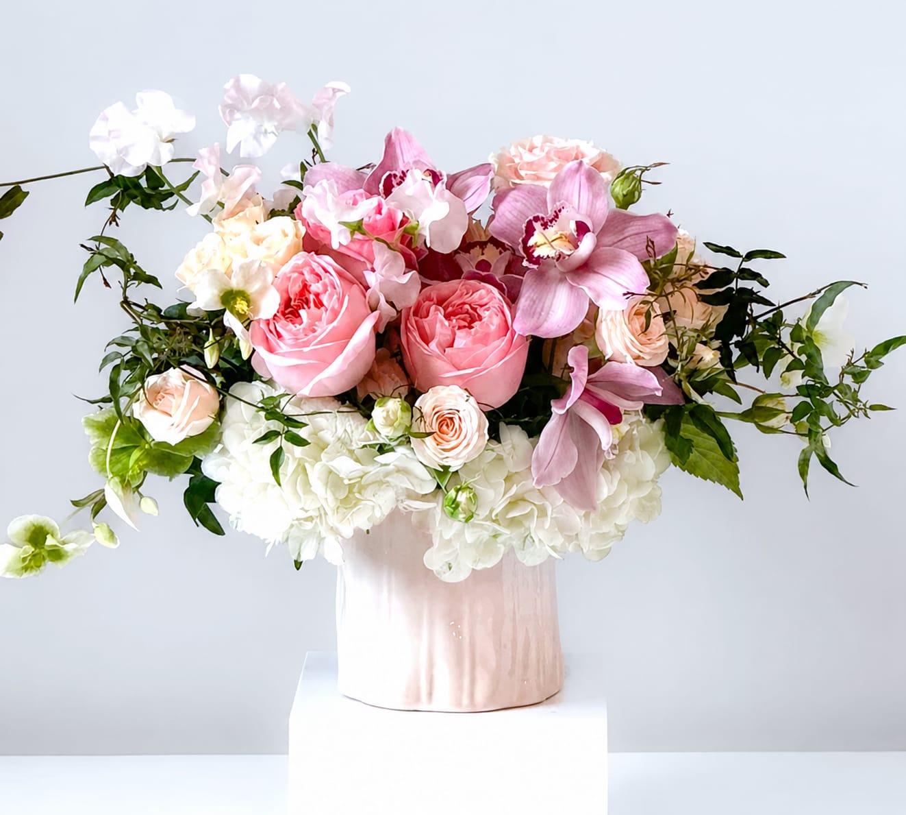 Blushing Hearts - A gorgeously lush arrangement featuring soft blush garden roses, hydrangeas, and New Zealand cymbidium orchids in a white ceramic vase. 