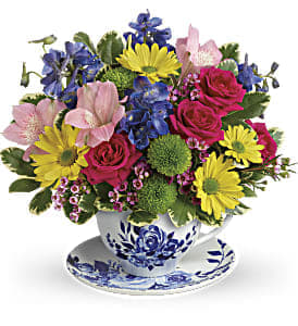 Colorful mixed bouquet of pink roses, yellow daisies and blue flowers arranged in a blue-and-white teacup.
