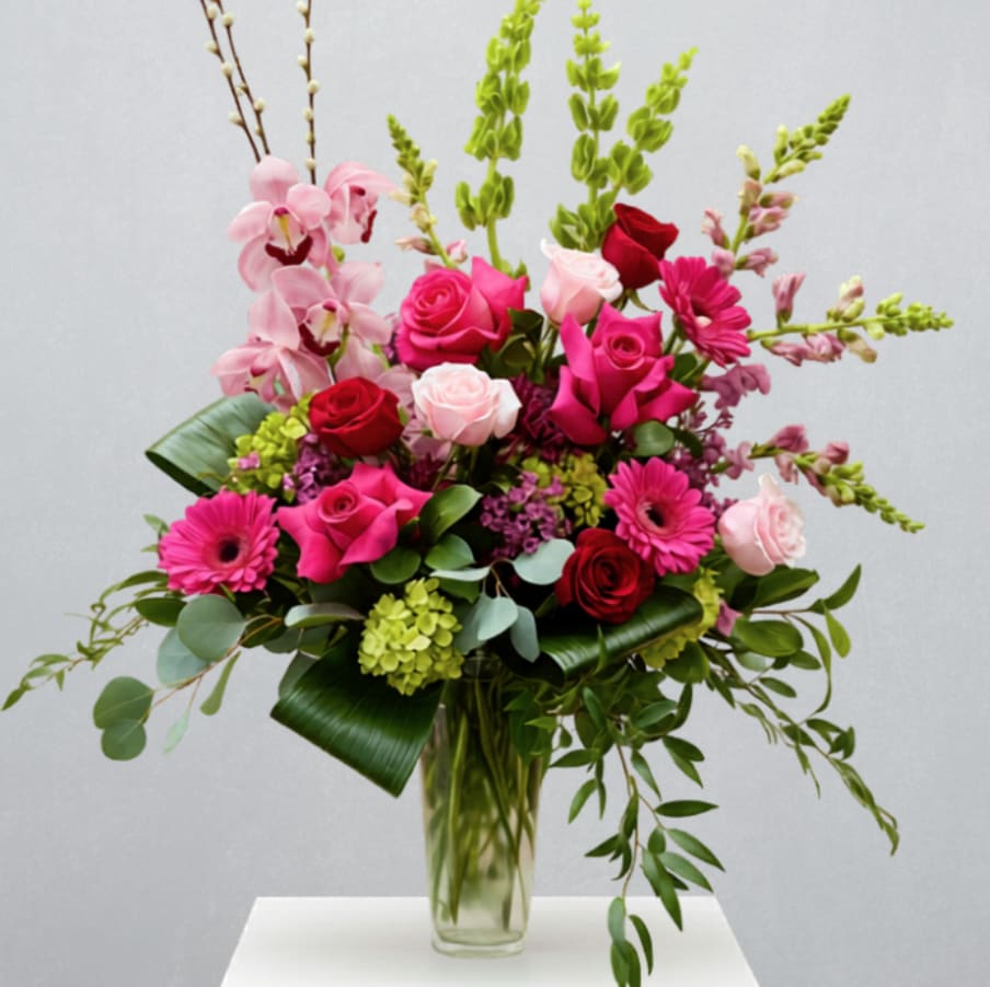Iconic Blooms - EVERYDAY COLLECTION | LOCAL DELIVERY ONLY  A show-stopping arrangement designed in a tall Giselle vase. This elevated piece features bright, romantic hues of roses, mojito hydrangea, a cymbidium orchid stem, gerbera daisies, mums, bells of ireland, snap dragons, pussy willow, wax flower, and lush greenery- created to make a bold, unforgettable statement of love. 