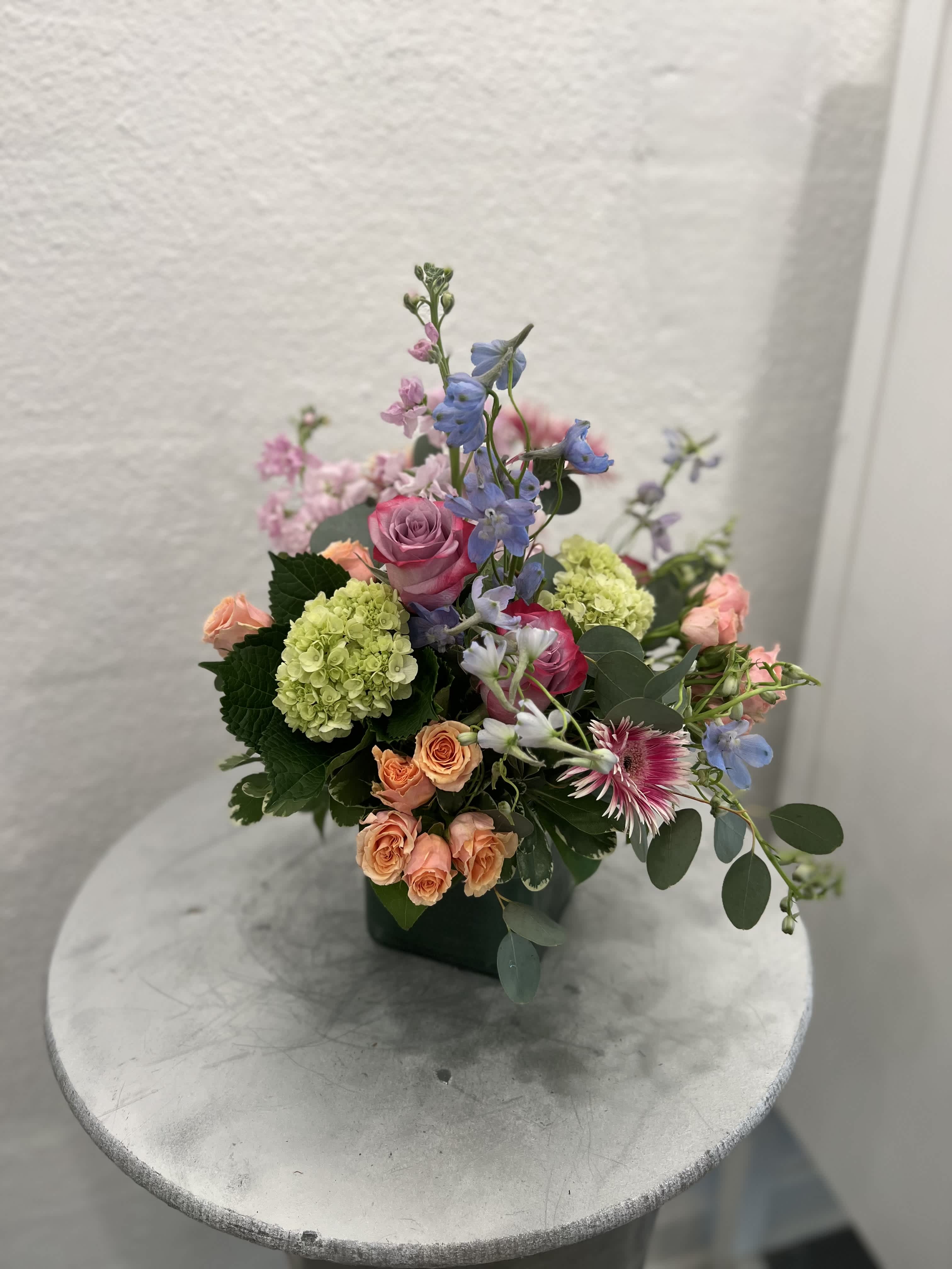 Pastel arrangement of pink and peach roses, green hydrangea and blue blooms in a green square vase