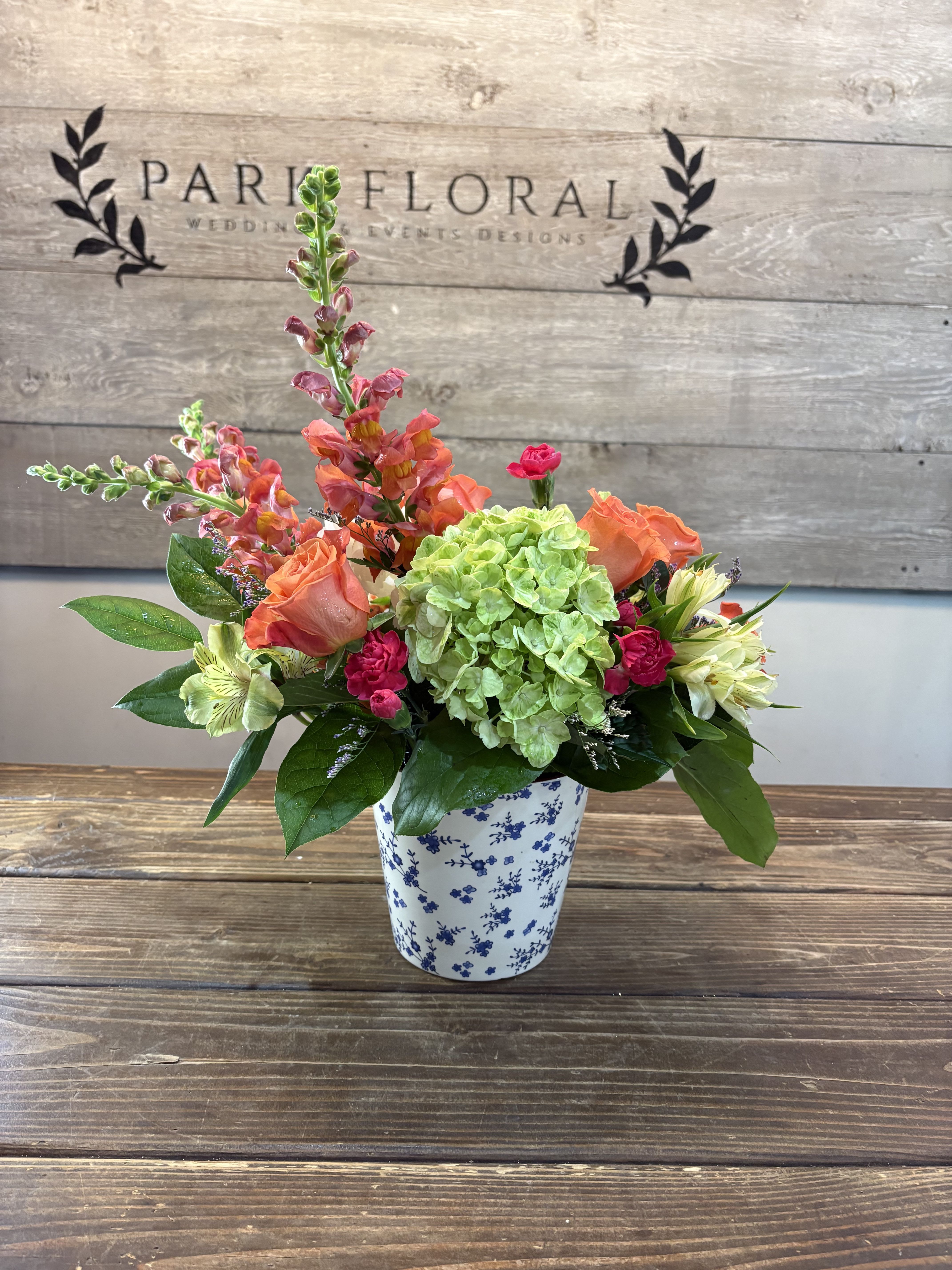 Summer Breeze - A lively cluster of coral-orange roses and bright green hydrangea makes an immediate sunny impression-perfect as a birthday bouquet or a "just because" surprise that brings a fresh spring feeling indoors. This flower arrangement blends pink snapdragons and hot pink carnations with pale alstroemeria for a mix of height, texture, and cheerful color that will make the person you're thinking of feel noticed and celebrated.  Compact and rounded with a few taller spikes for movement, the arrangement is presented in a white ceramic pot with blue floral decoration that gives it a cottage-style, homey charm. Blooms include alstroemeria, carnations, hydrangeas, roses, and snapdragons in shades of pink, white, green, and orange-an uplifting palette that reads both lively and balanced.  Ideal for birthdays, thank-you gifts, springtime decorating, or an everyday pick-me-up, this piece suits a table centerpiece, entryway, or desk. The ceramic pot and vase presentation makes it gift-ready; to keep blooms looking their best, place in a cool spot away from direct sun and refresh the water if transferring to a vase.