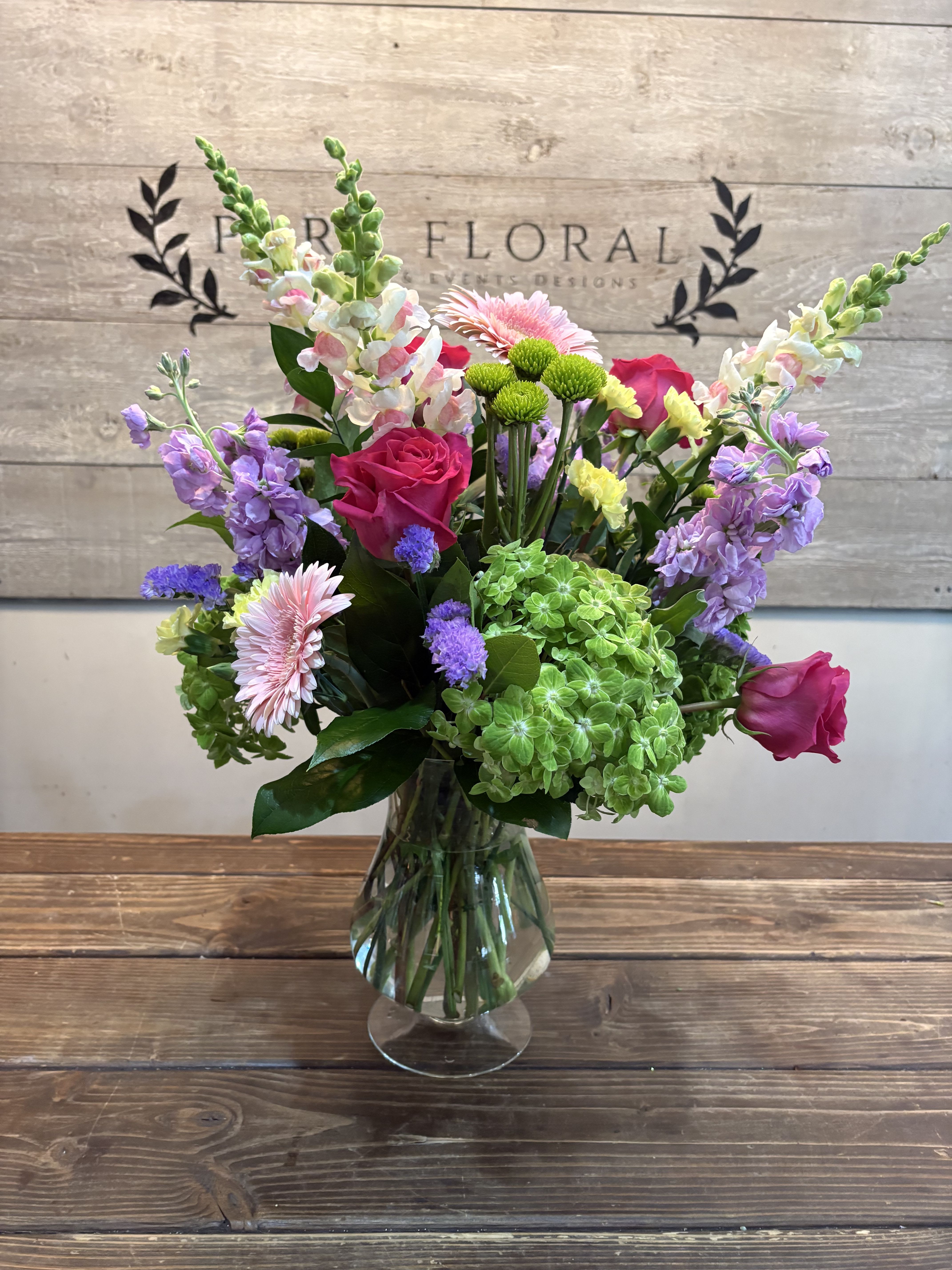 Secret Garden - Bright pink roses and vivid gerbera daisies anchor this Secret Garden flower arrangement, a cheerful flower arrangement perfect for birthdays, thank-you gifts, or spring celebrations. Lavender snapdragon spikes and clusters of green hydrangea add height and garden-fresh texture while soft white and pale pink accents keep the palette light and uplifting - the kind of bouquet that makes the person you're thinking of feel seen and celebrated.  The mix includes roses, gerbera daisies, hydrangeas, snapdragons, chrysanthemums, carnations, and delicate filler blooms in pink, purple, green, and white. Upright snapdragon stems contrast with rounded hydrangea and daisy heads to create a full, airy composition with a polished, garden-style look. Arranged in a clear glass vase, it presents cleanly on a table or desk and arrives ready to display.  Ideal for sending warm wishes or brightening someone's day, Secret Garden suits recipients who love fresh, colorful bouquets with a lively yet refined feel. The compact, upright design fits well on mantels, side tables, or office desks - order for same-week delivery to make sure it arrives at peak freshness.