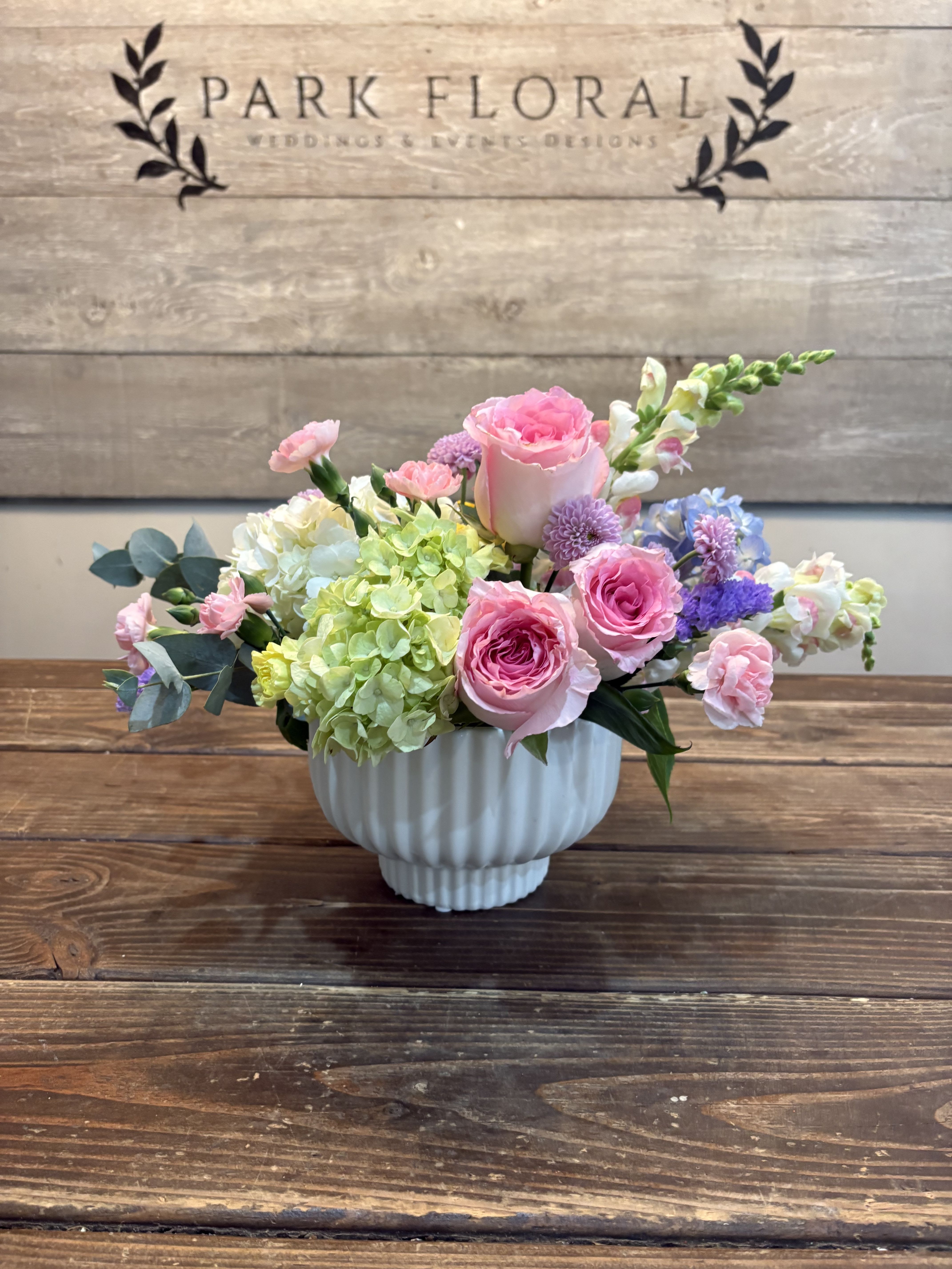 Cherry Blossom - Soft blush roses and pale green hydrangea create a gentle, garden-fresh centerpiece in this Cherry Blossom flower arrangement - perfect as a birthday bouquet, anniversary surprise, or a thoughtful thank-you gift. The rounded, low profile and pastel palette (pink, white, lavender, yellow-green) make the recipient feel genuinely cherished: romantic without being formal, cheerful without being over the top.  Layered textures - full hydrangea clusters, velvet roses, small pink carnations, lavender button asters, snapdragon spikes, and delicate blue-white accents - give the design depth and a hand-gathered look. The flowers are arranged compactly in a ribbed white ceramic vase that reads classic and modern at once and keeps the overall composition neat on a table or desk.  Designed to brighten everyday moments or celebrate springtime occasions, this arrangement suits someone who loves soft color and garden-inspired style. The compact size fits well on mantels or as a centerpiece; to keep blooms looking their best, change vase water regularly and trim stems if needed before arranging.