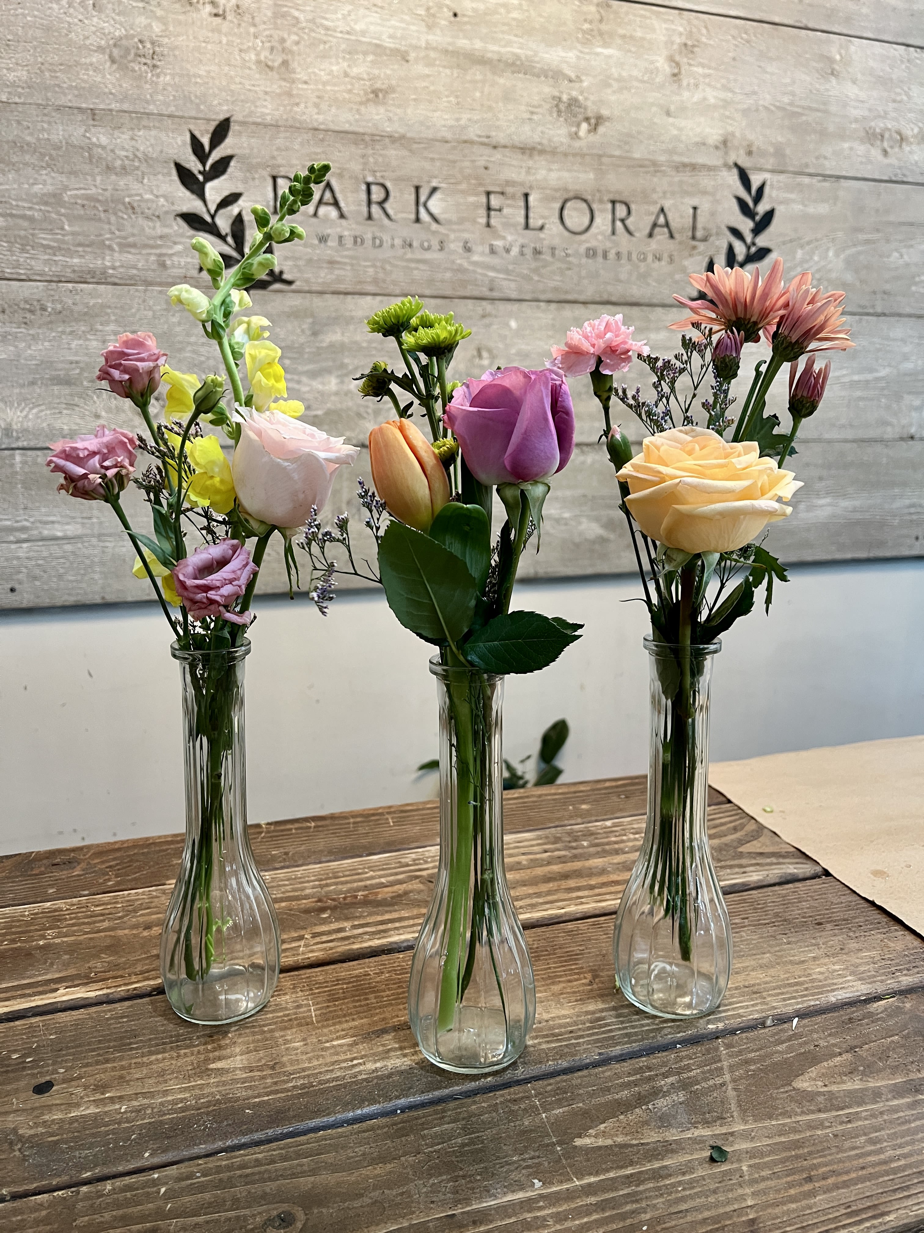 Bud Vase Arrangement - These petite arrangements pair roses, tulips, carnations, snapdragons, and small green chrysanthemums with airy filler stems for height and texture, arranged in narrow clear glass bud vases. Colors run from blush pinks and lavenders to pale peach, soft yellow, white and a touch of orange, giving each vase its own personality while remaining a coordinated set. The scale is intentionally small and casual-each vase is a single-flower focal with complementary accents-so they fit easily on a bedside table, desk, or windowsill.  Choose this set when you want a simple, thoughtful gift that looks hand-delivered: ideal for everyday gifting, a quick birthday surprise, or a bright "thank you." The clear glass bud vases arrive ready to display; refresh water every 2-3 days and trim stems slightly for best longevity.