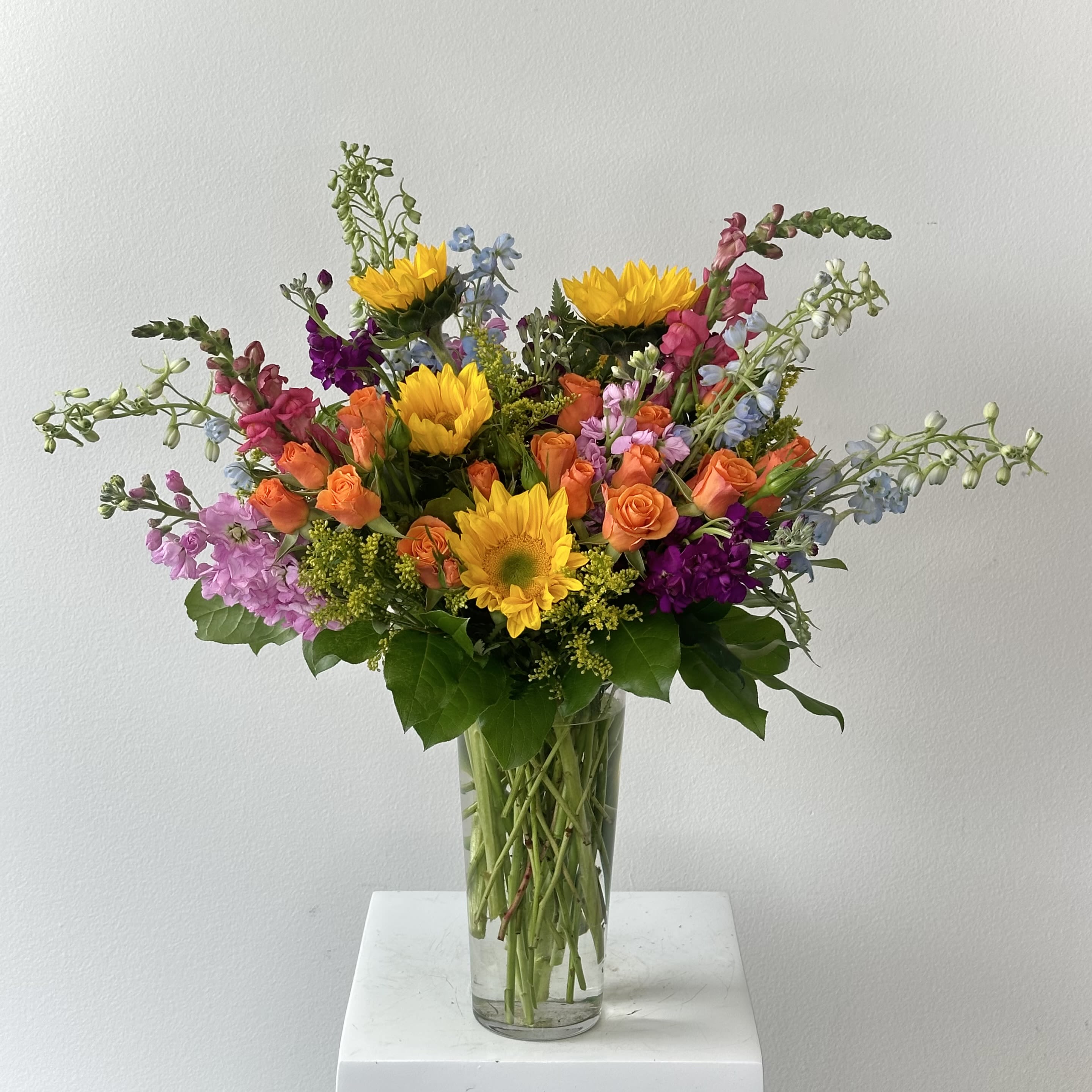 Full Nest Bouquet - Bask in a glow of affection with Irvine Village Flowers’ full nest bouquet—a sunlit embrace that wraps you in love from every angle! This radiant creation blooms with golden sunflowers, their cheerful faces beaming like the sun itself, alongside tender roses that hum a sweet melody of the heart. Delphiniums rise tall, their spires of blue and purple adding a touch of skyward magic. Together, they nestle into a full, lush arrangement, a warm hug of flowers that radiates joy and devotion.  Each bloom weaves a tale of warmth: sunflowers burst with happiness and adoration, their bright petals a shout of love that lights up the room. Roses, soft and velvety, whisper romance and care, while delphiniums stretch upward, threading the bouquet with hope and grace. From Irvine Village Flowers, this bouquet is a circle of affection, a gift that surrounds you with beauty. Stop by our shop or order online, and let our seamless delivery bring this radiant delight to your doorstep, fresh and ready to fill your world with love from every direction!
