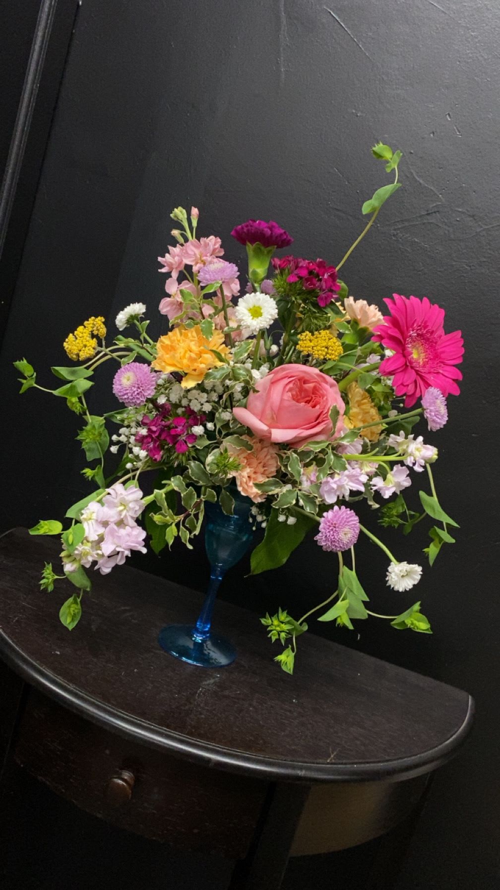 Summer Meadow - A large pink rose anchors this playful garden-style bouquet, a birthday bouquet or just-because arrangement that reads like a sunny spring moment. The mix of pink, purple, yellow, white and peach blooms-featuring roses, a bright magenta gerbera daisy, carnations, daisies, chrysanthemums and alstroemeria-creates an airy, carefree silhouette that will make the person you're thinking of feel celebrated and seen.  Set in a cobalt-blue glass vase, the composition uses visible branching stems and scattered blossoms to add height and movement; the magenta gerbera provides a vivid focal point while clustered flowers and softer roses soften the overall texture. Sized to sit on a table or desk, this loose, informal design brings cheerful color without feeling fussy.  Perfect for springtime birthdays, congratulations, or a spontaneous pick-me-up, this arrangement arrives ready to display in the included blue glass vase. To keep the flowers looking fresh, change the water every couple of days, trim stems at an angle, and remove any foliage below the waterline.